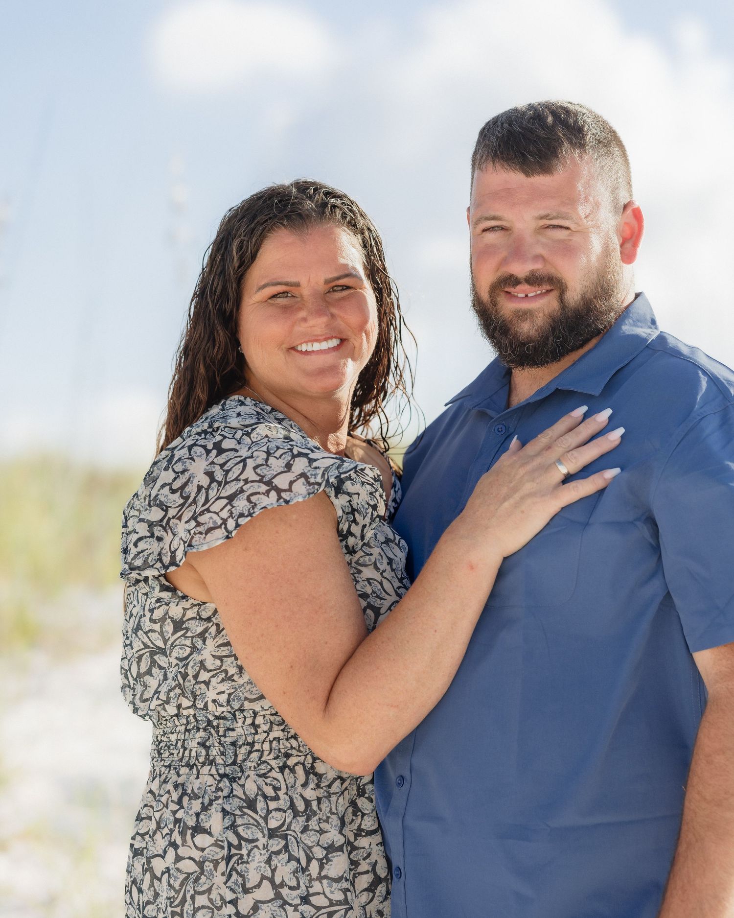 A smiling couple poses together on a sandy beach, the woman in a floral dress and the man in a blue shirt.