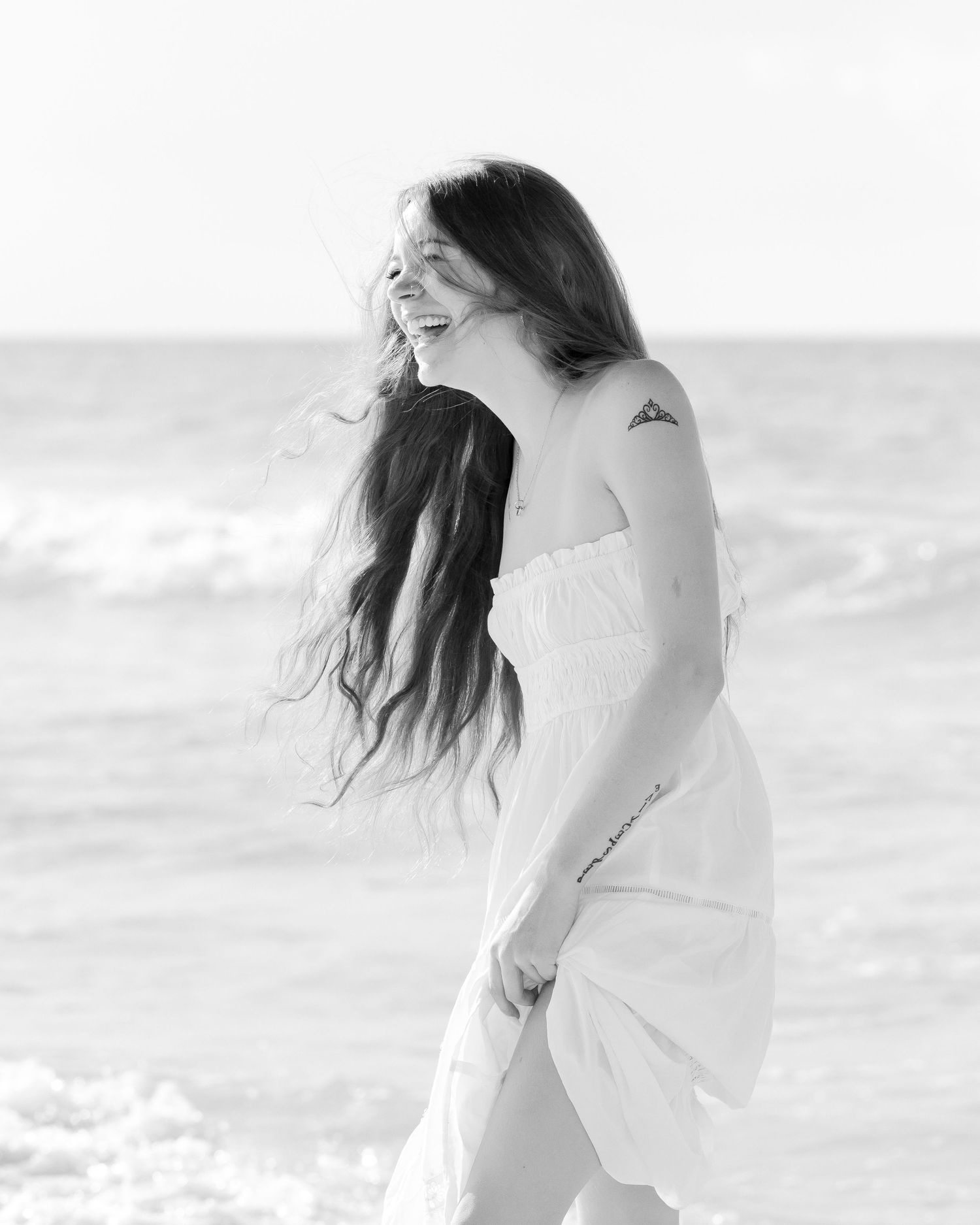 Black and white photo of a smiling woman in a white off-shoulder dress on a beach with ocean waves behind her.