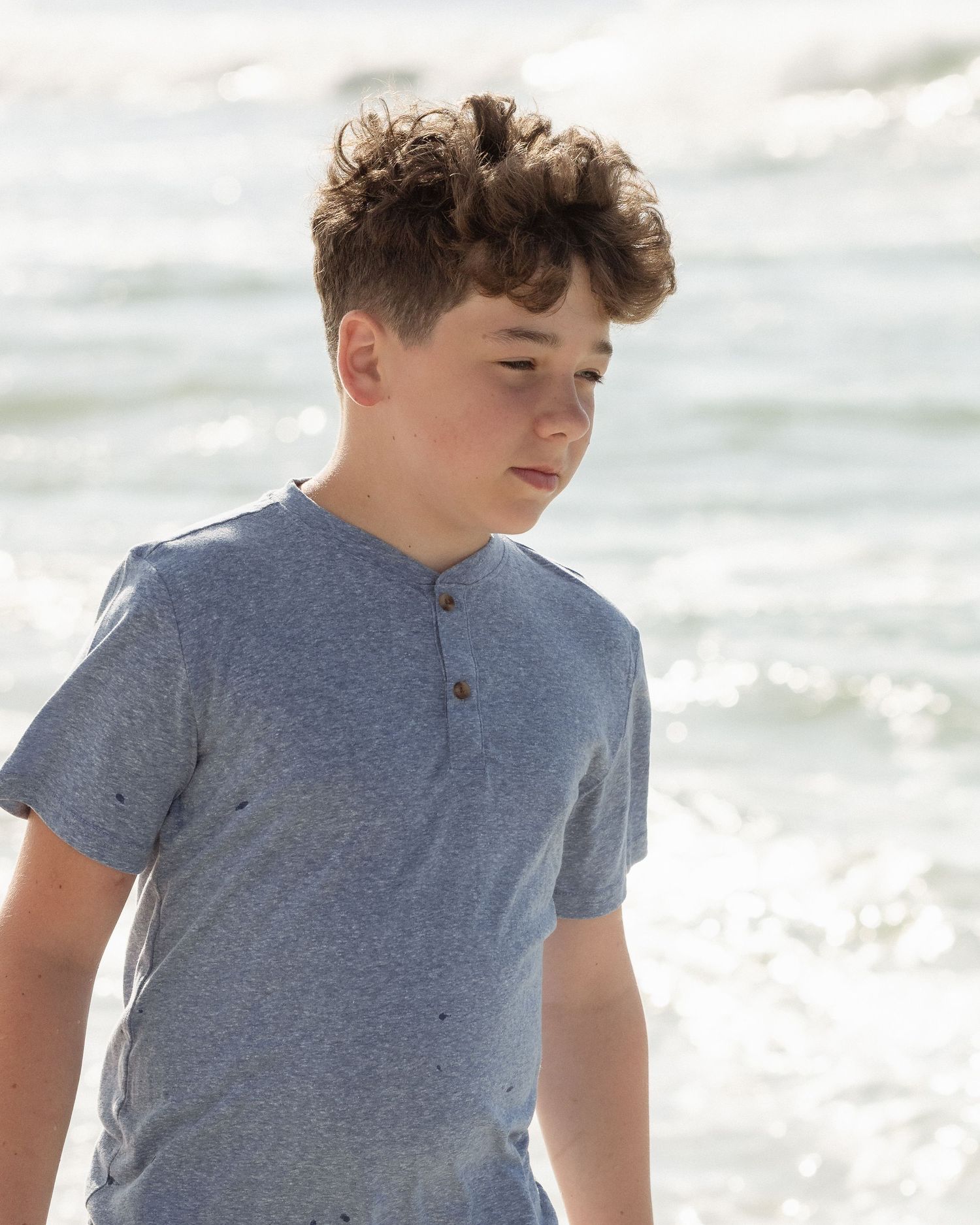 A teenage boy with curly hair wearing a blue henley shirt stands at the beach with ocean waves behind him.