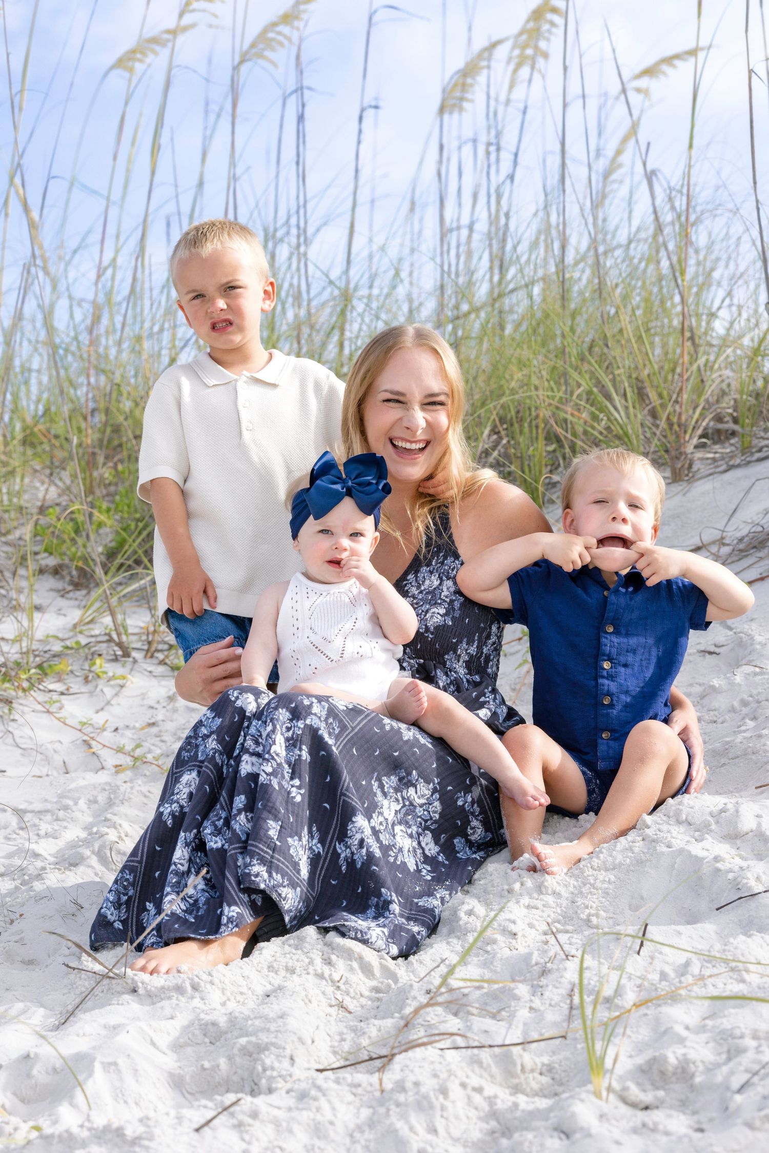 A smiling mother sits on white sand beach dunes with her three young children, wearing coordinated navy blue outfits.
