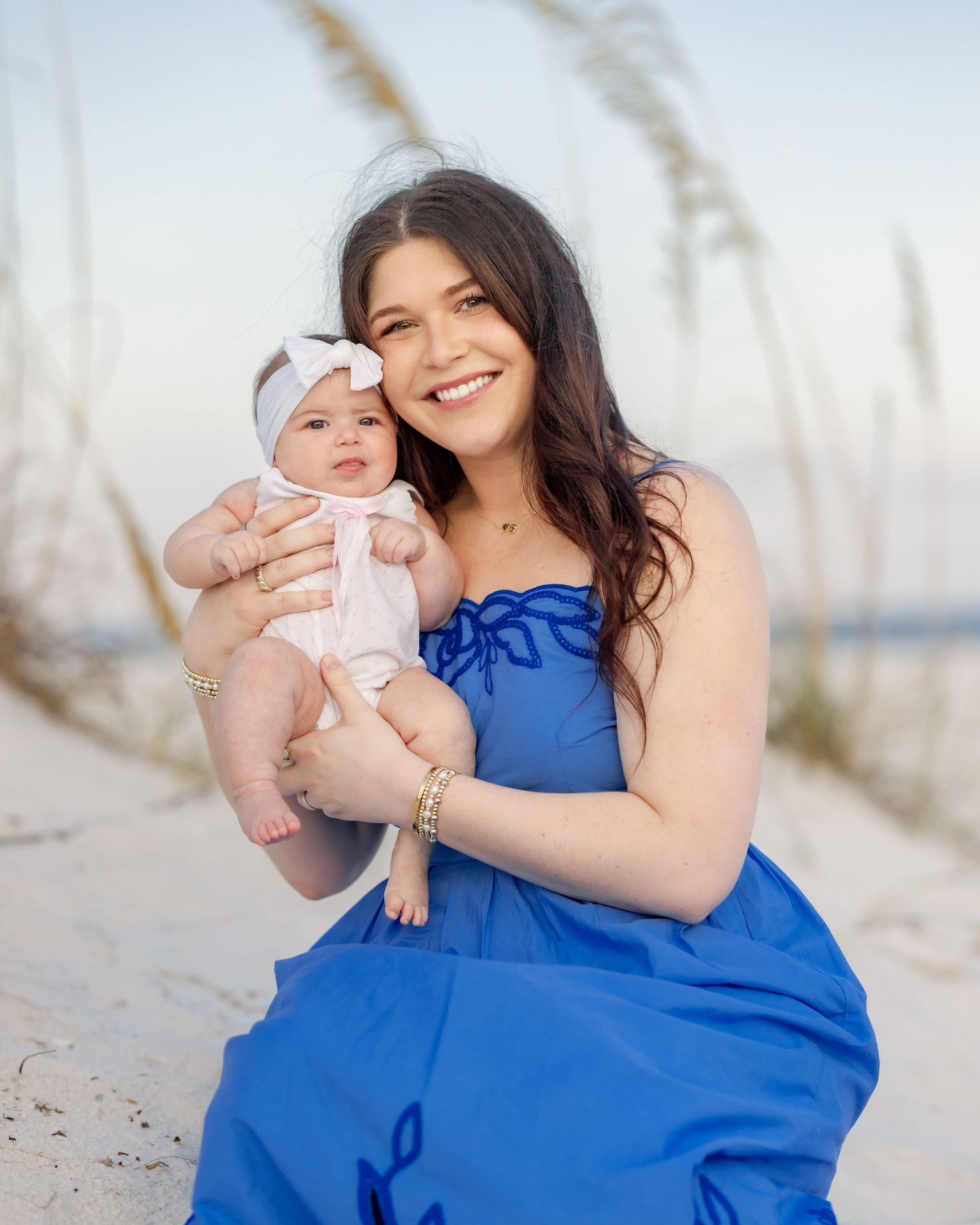 Smiling brunette woman in blue dress holding adorable baby with bow headband on beach.