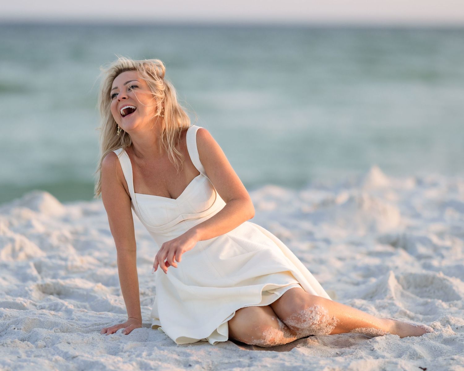 A cheerful blonde woman in a white dress sits laughing on white sand beach.