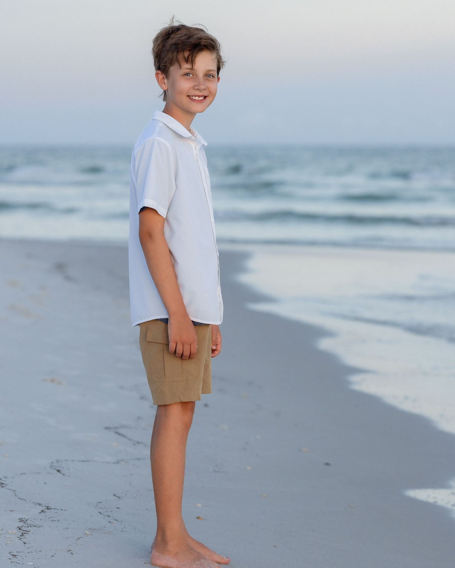 A smiling boy in a white shirt and khaki shorts stands barefoot on a sandy beach with ocean waves behind him.
