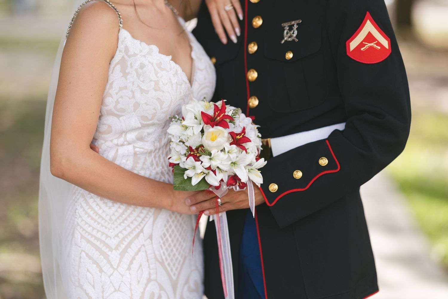 Marine in dress uniform stands beside bride in white lace gown holding red and white floral bouquet.
