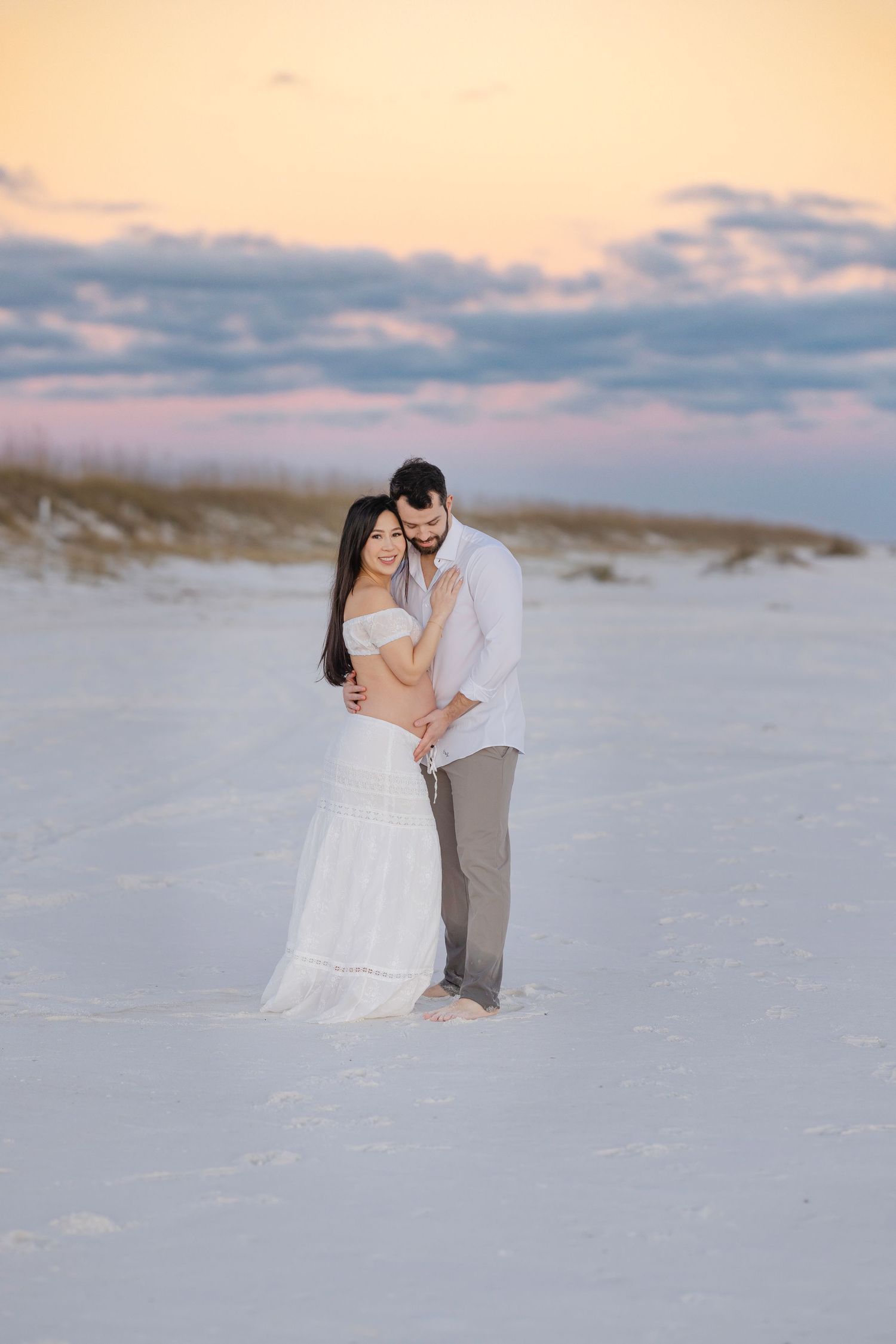 A romantic beach wedding photo session with a couple embracing on white sand dunes at sunset.