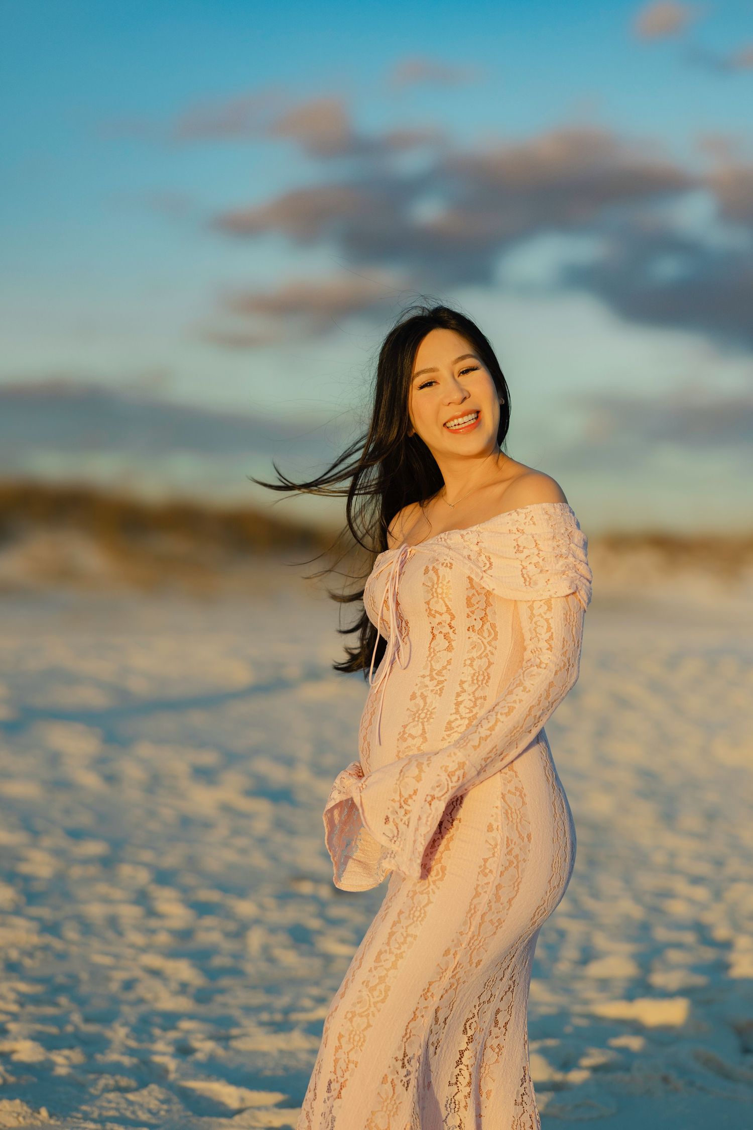 Woman in flowing pink lace dress poses on beach at golden hour with hair blowing in wind.
