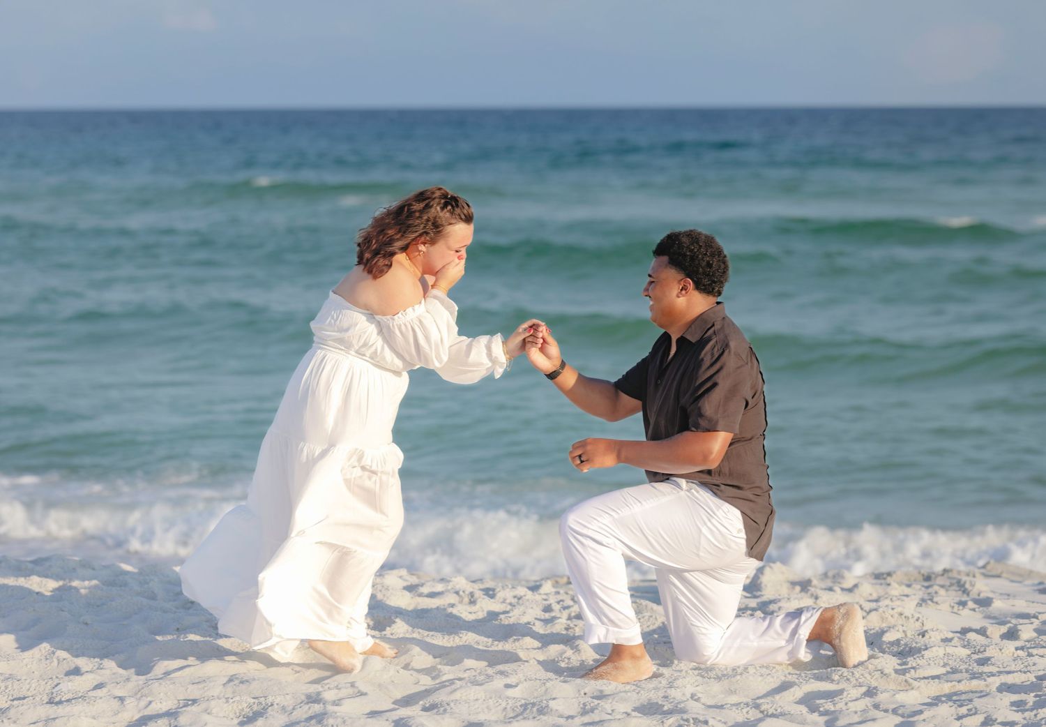 Man kneeling on sandy beach proposing to woman in white dress with ocean waves in background.