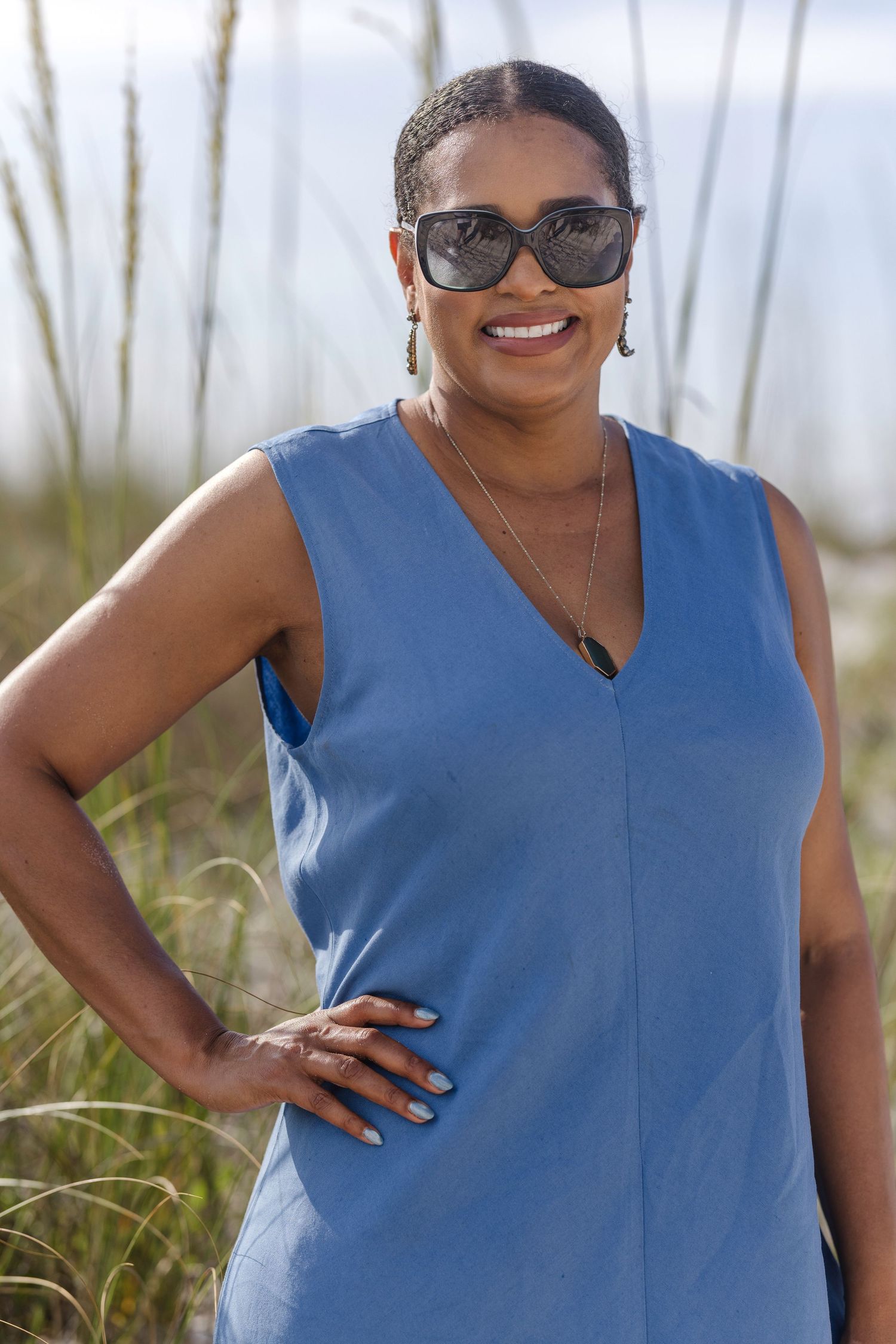 Smiling woman in blue sleeveless dress and sunglasses posing on a beach with sea grass in background.