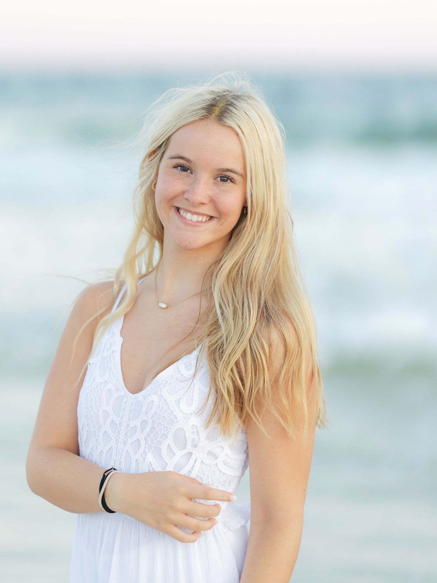 Smiling blonde teenage girl in white dress posing at beach for senior portrait photo.
