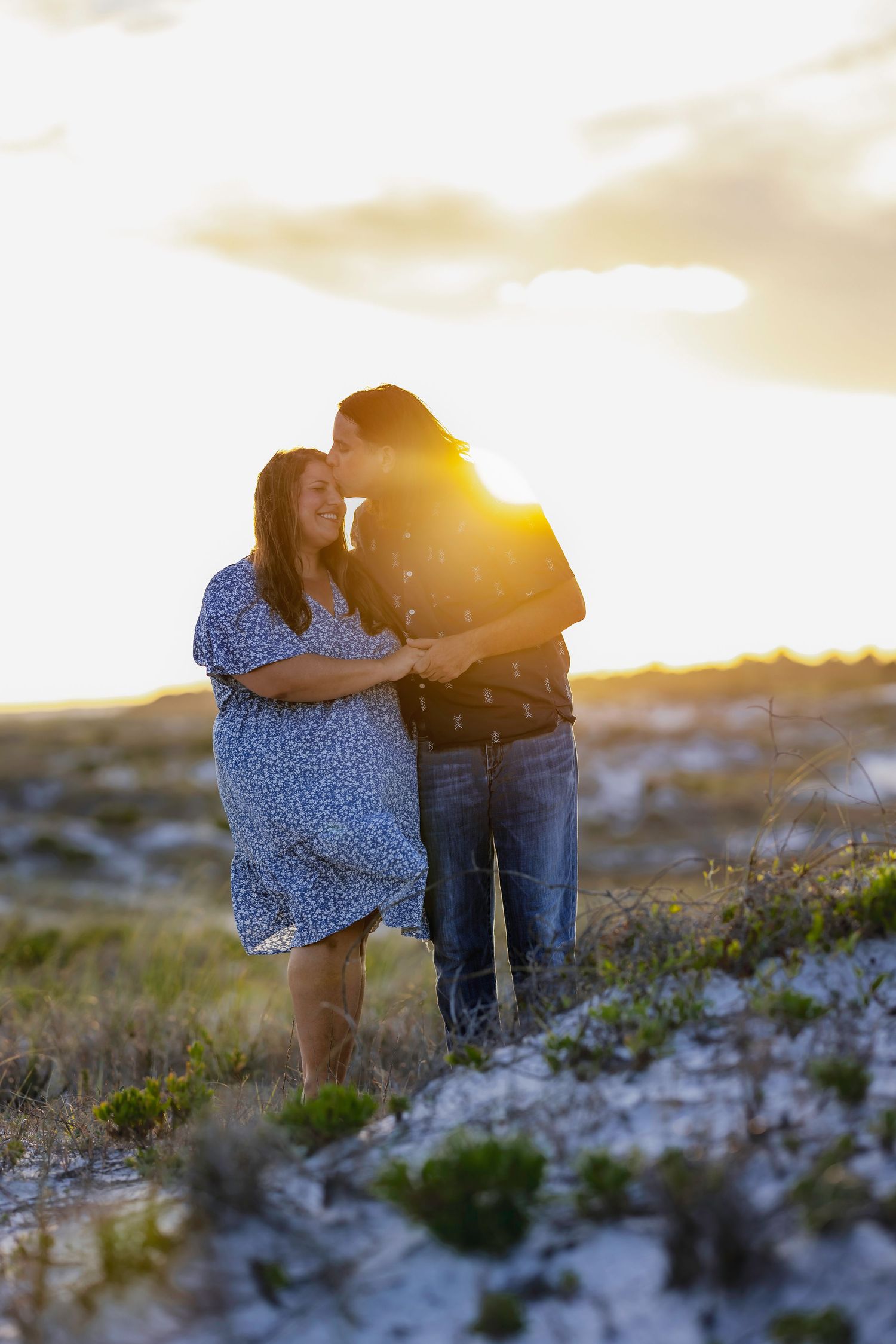 Couple embracing at golden sunset on sandy beach, woman in blue floral dress, backlit by warm light.
