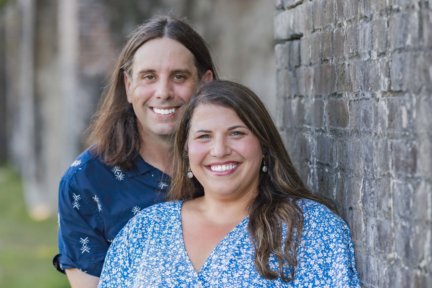 Smiling couple posing outdoors near a brick wall, man with long hair in blue shirt, woman in floral dress.