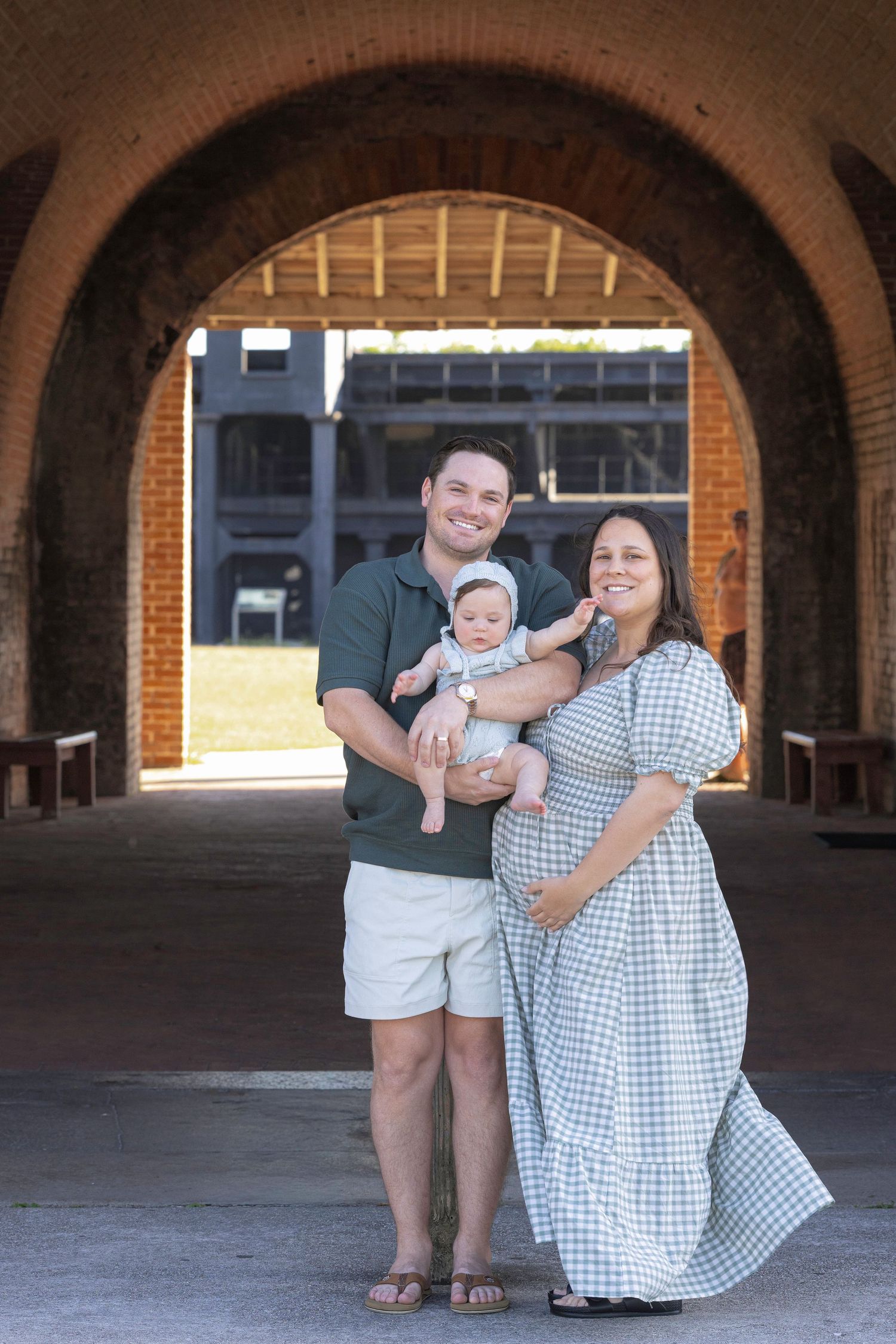 Smiling family with pregnant mother in gingham dress and father holding baby under brick archway.