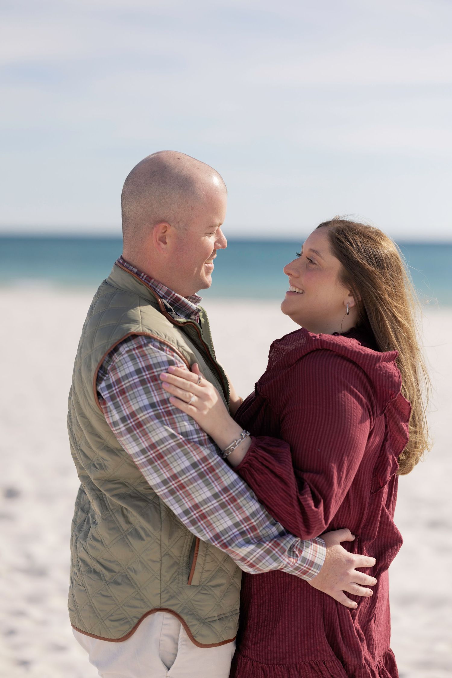 Happy couple hugging and smiling at each other on a sunny beach, man in green vest and woman in burgundy dress.