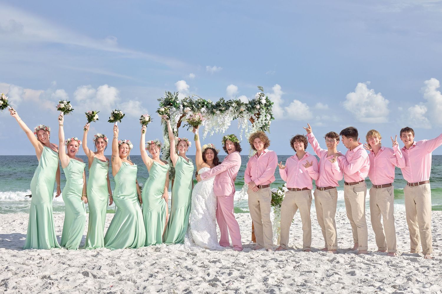 Beach wedding party in mint bridesmaid dresses and pink groomsmen shirts celebrating joyfully on a sunny sandy shore.