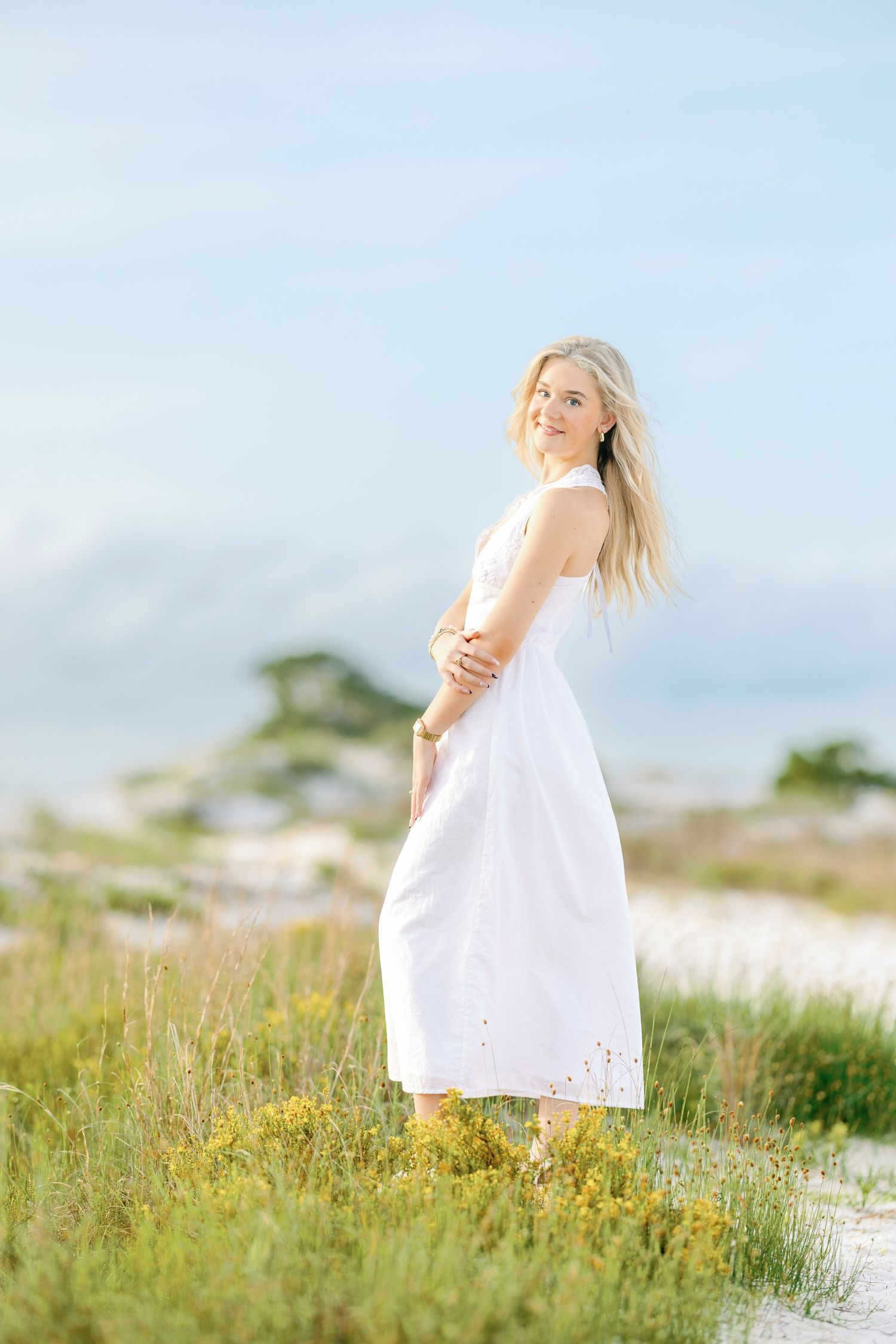 Blonde woman in white midi dress standing among beach dunes and wildflowers under a soft blue sky.
