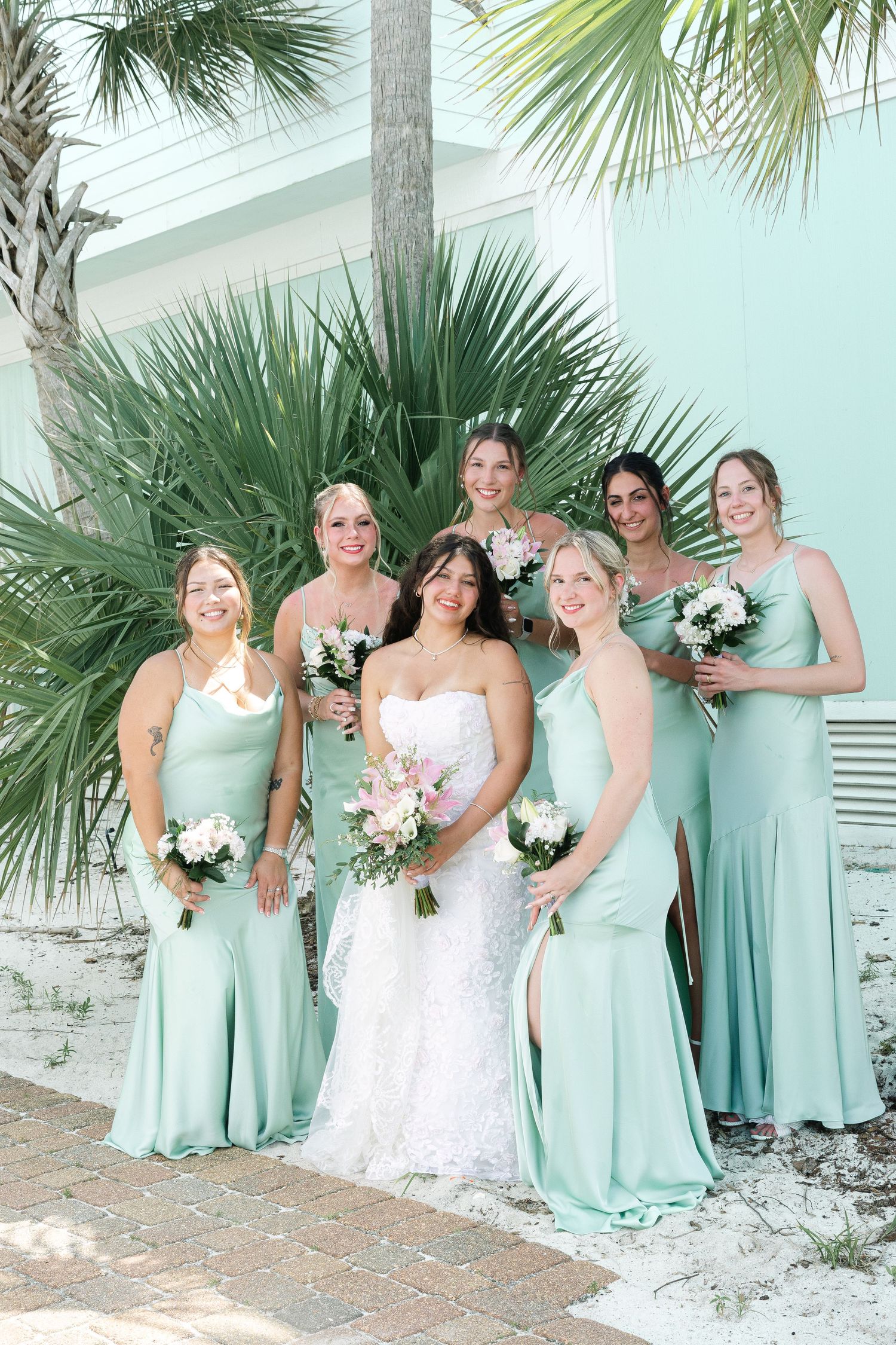 Bride in white lace gown poses with five bridesmaids in mint green dresses, holding bouquets outdoors near palm trees.
