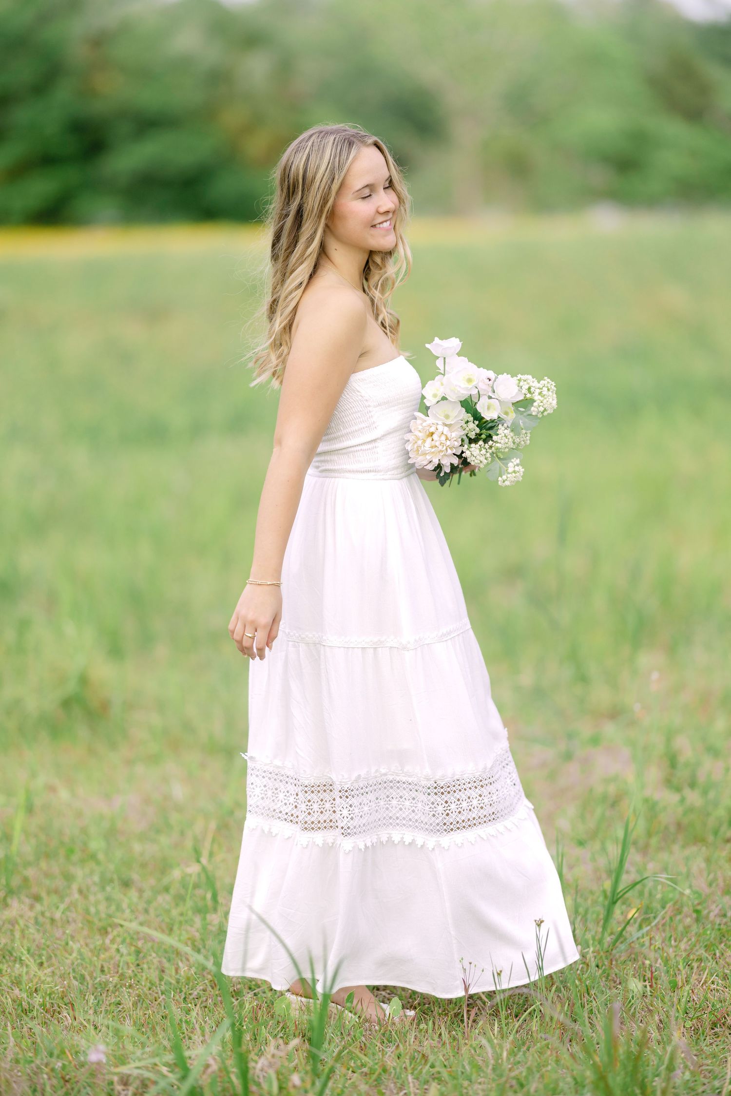 Young woman in white strapless maxi dress holding bouquet of pink and white flowers in a green meadow.