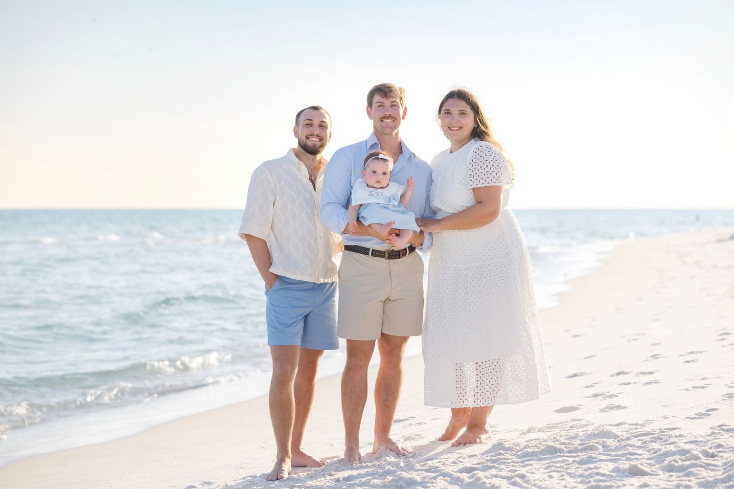 Family wearing white and blue outfits walking and posing together on a sunny beach during golden hour.