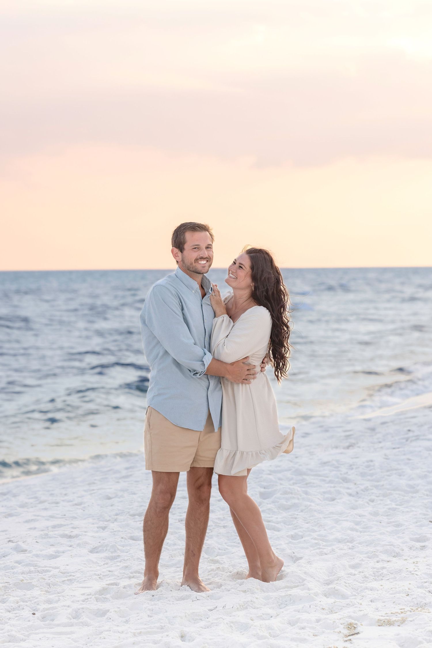 A romantic couple shares joyful moments on a sandy beach at sunset with waves crashing behind them.
