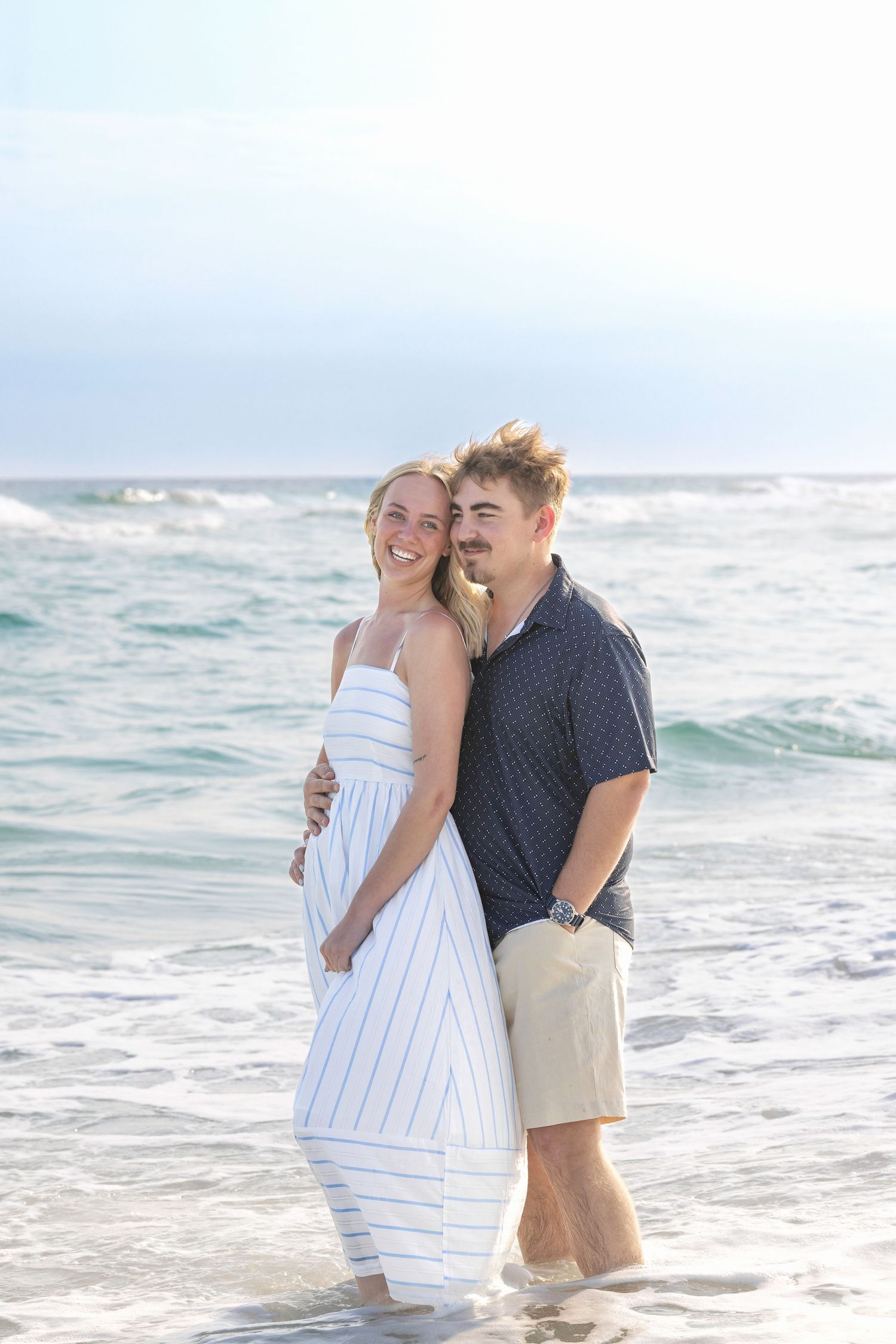 Happy couple embracing on a sunny beach, woman in white striped dress, man in navy shirt and khaki shorts, ocean waves behind.