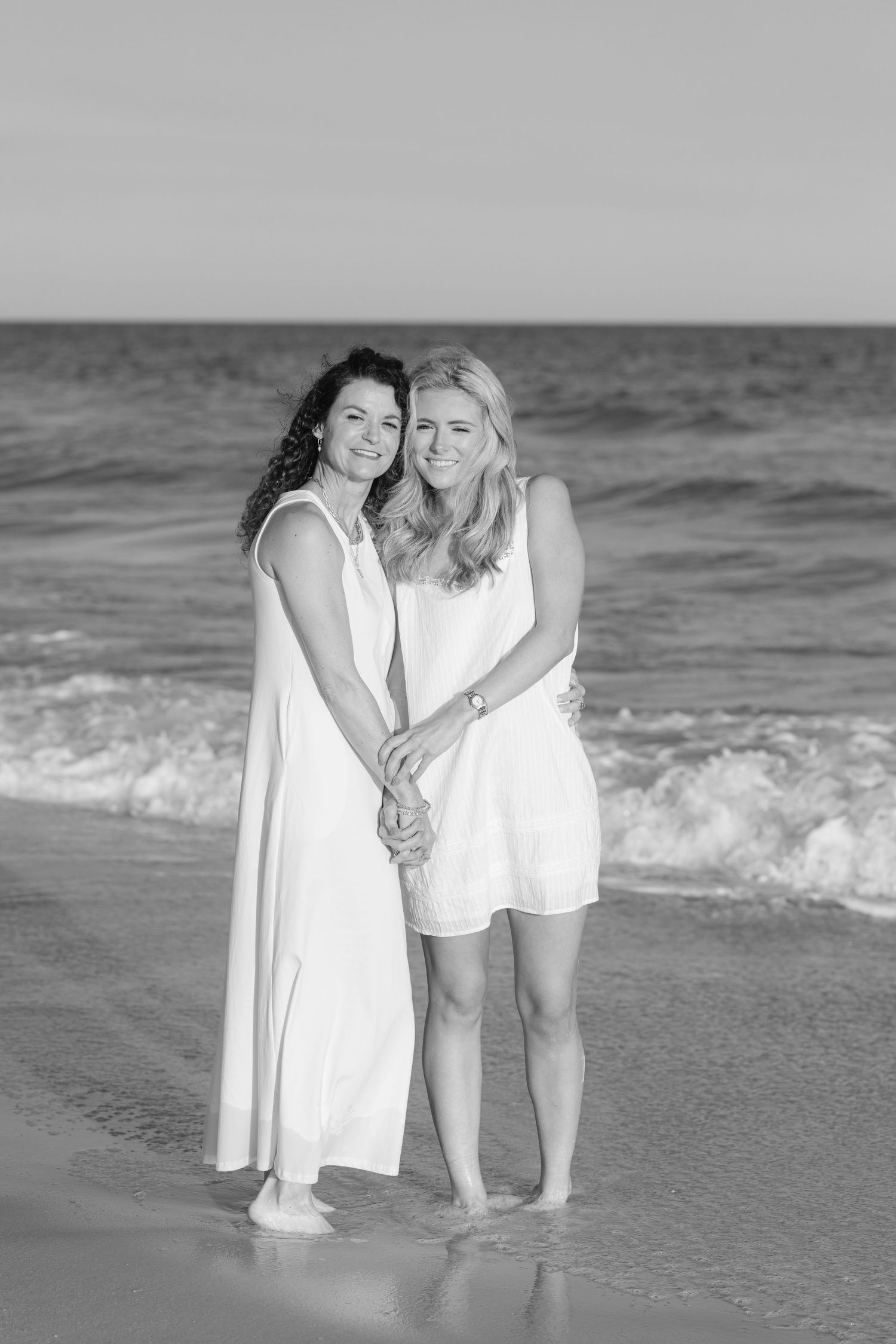 Two women in white dresses hold hands smiling on a beach shoreline in a black and white photo.