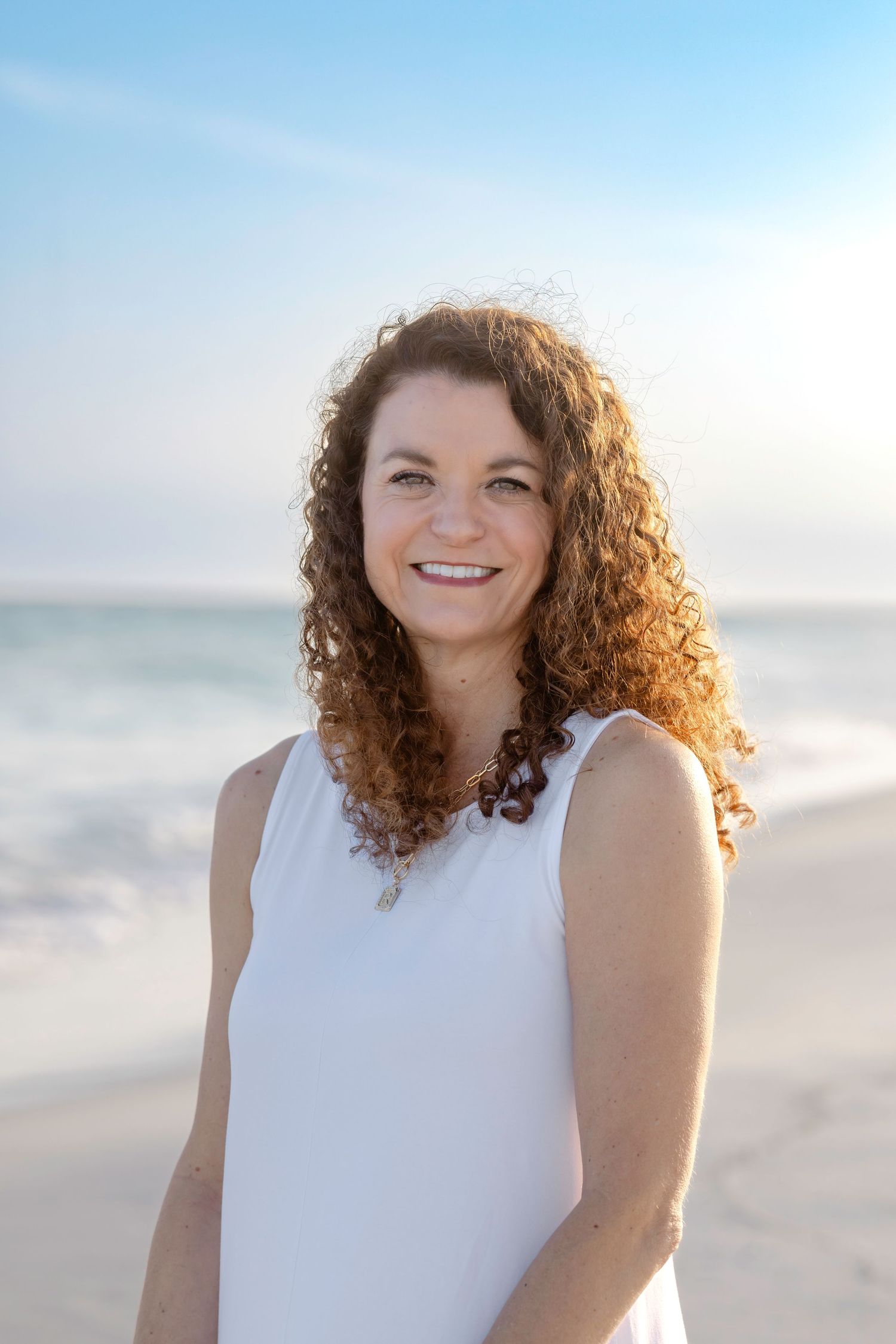 Smiling woman with curly auburn hair wearing a white sleeveless top, standing on a sunny beach with ocean waves behind her.