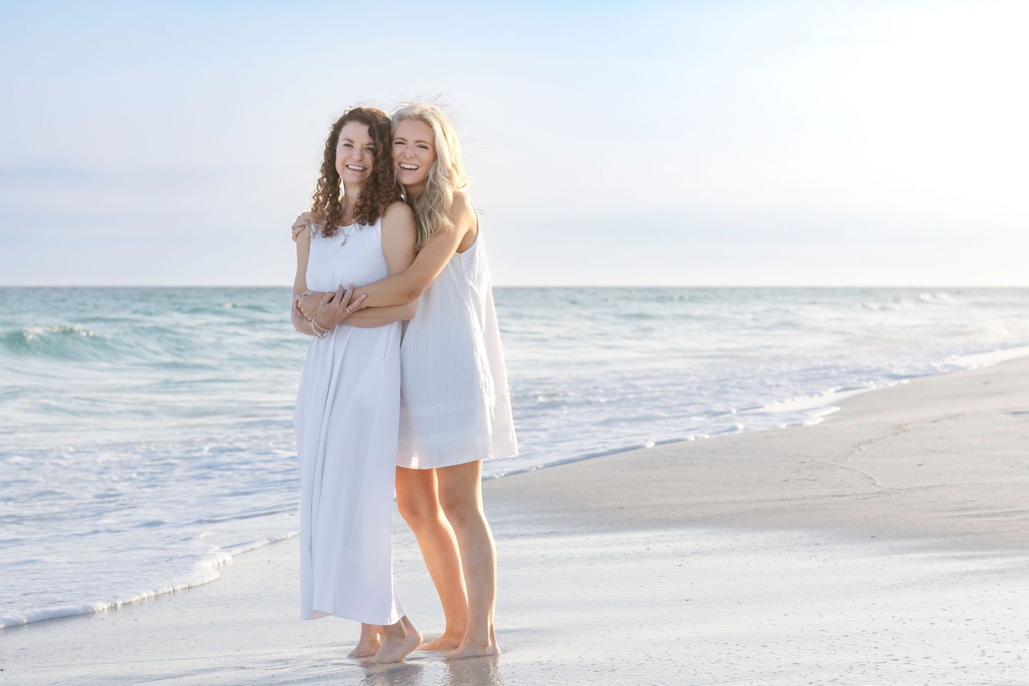 Two women in white dresses pose together on a sunny beach with turquoise waves behind them.