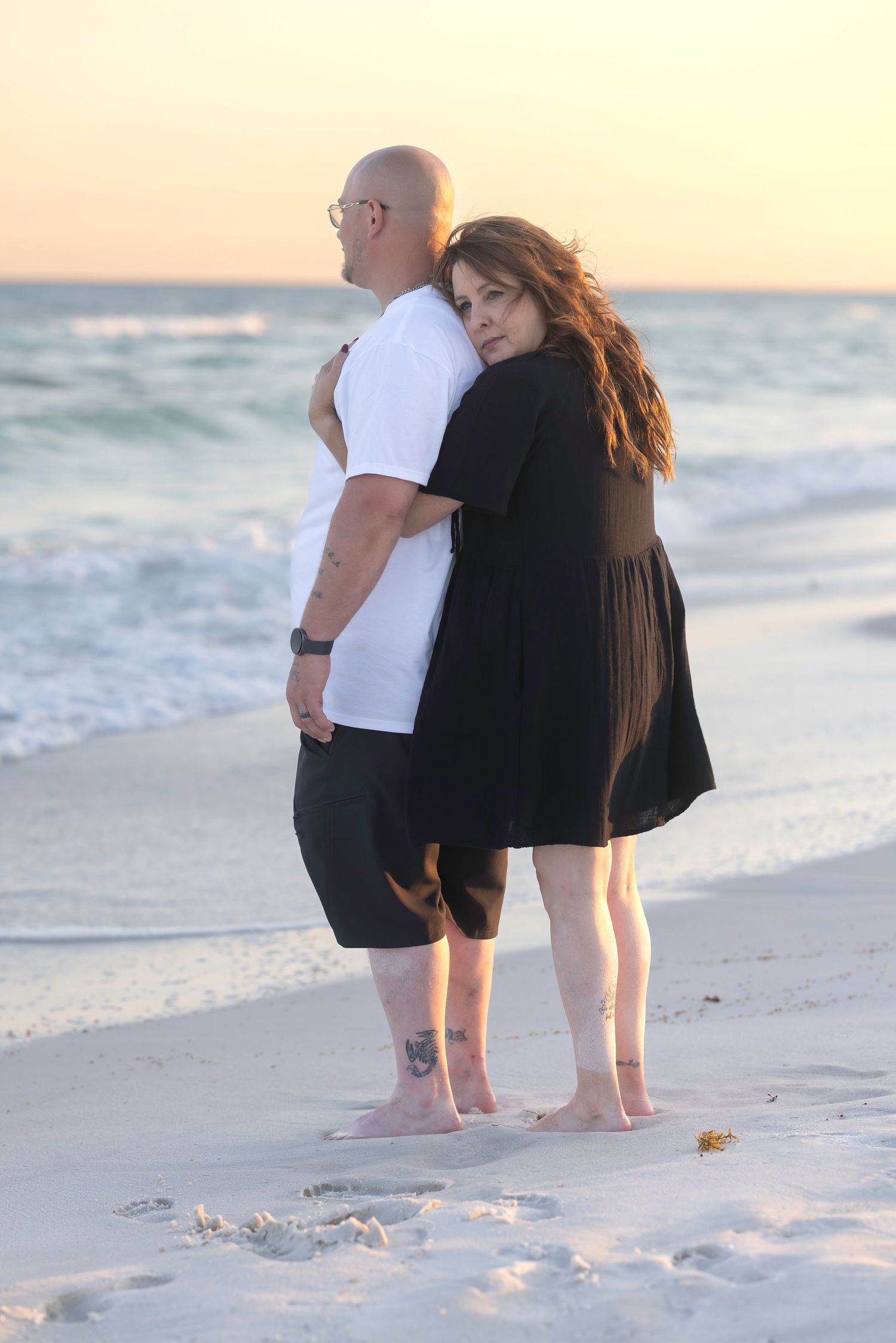 Couple standing back-to-back on sandy beach at sunset, both wearing black and white outfits near ocean waves.
