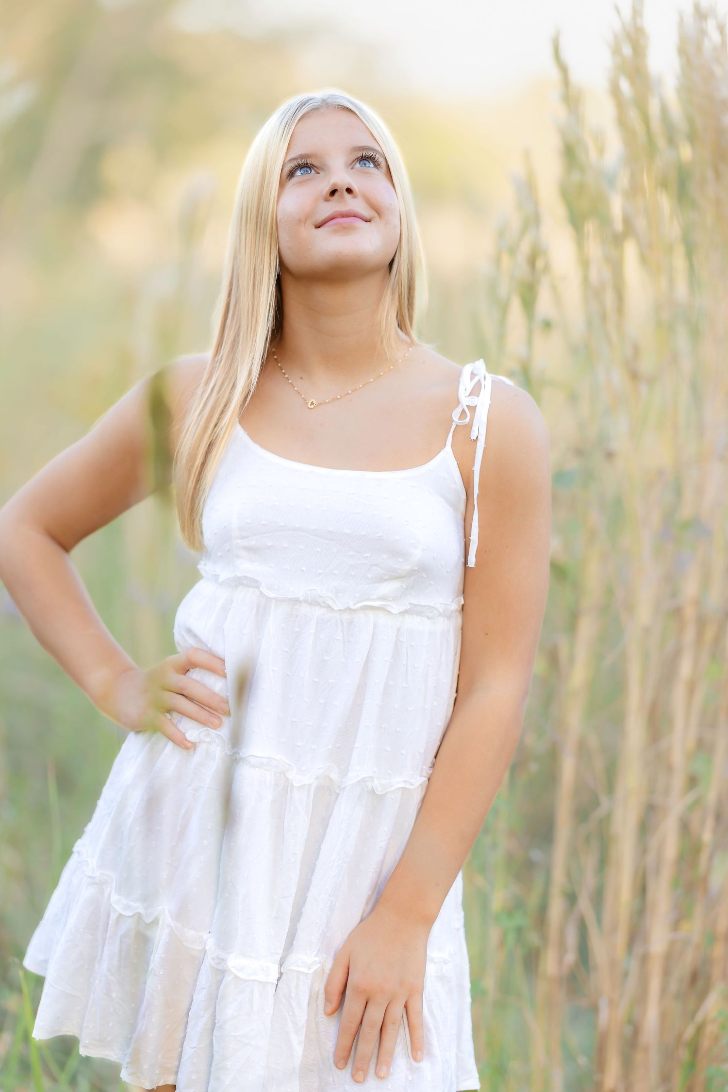 Young blonde woman in white sundress standing in tall grass field, looking up with a serene smile.