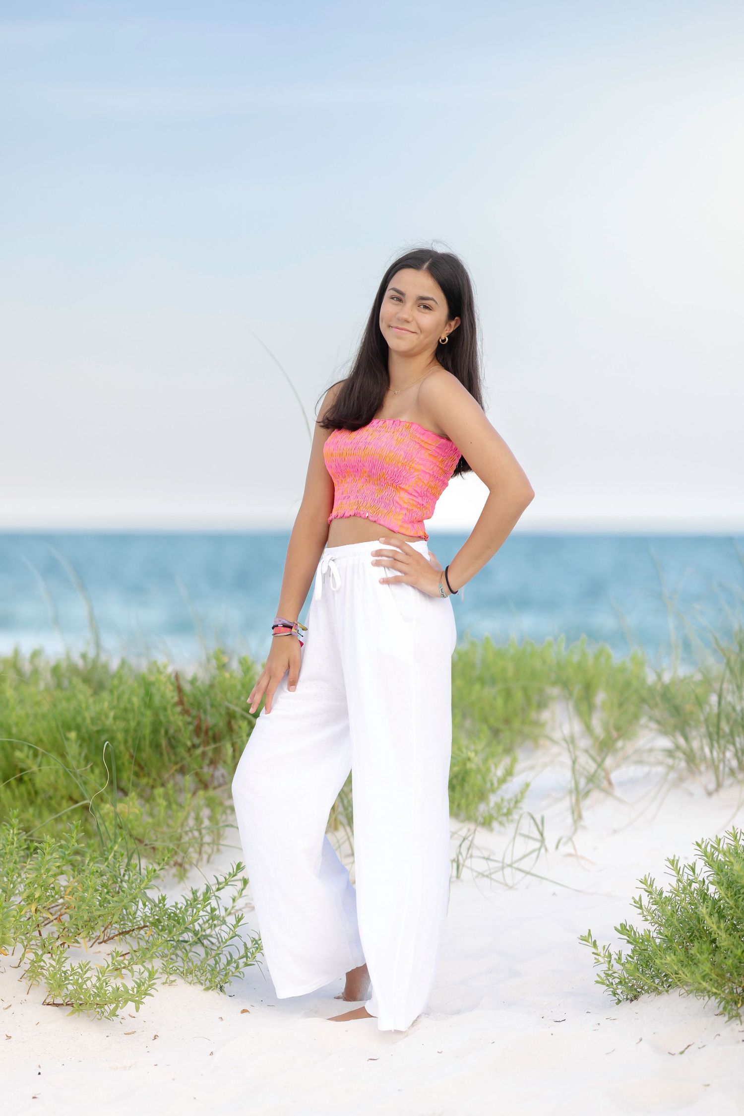 A person in a coral crop top and white palazzo pants poses on a sandy beach with ocean and grass in the background.