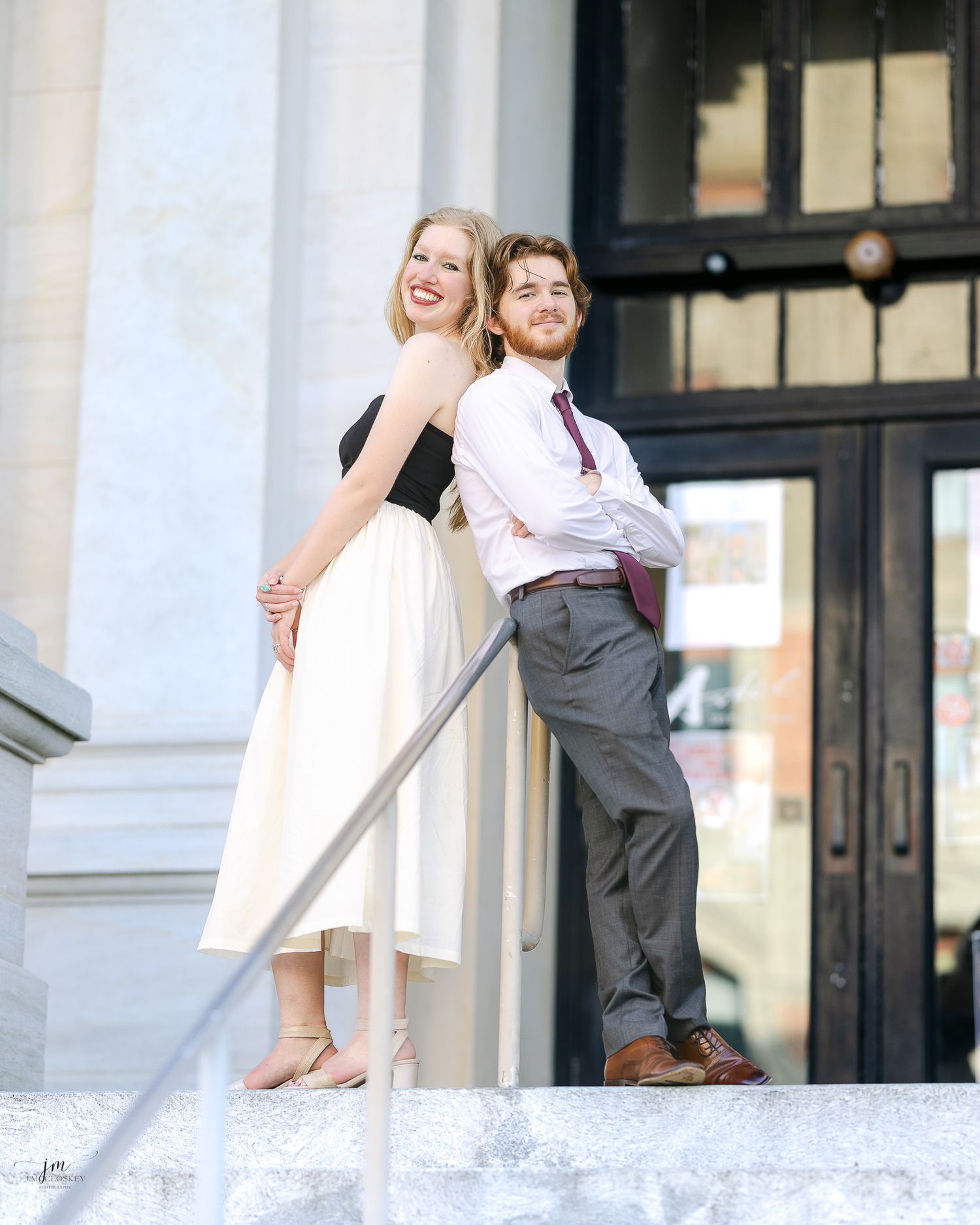 A couple in elegant attire stand back-to-back on white marble steps outside a building with ornate architecture.