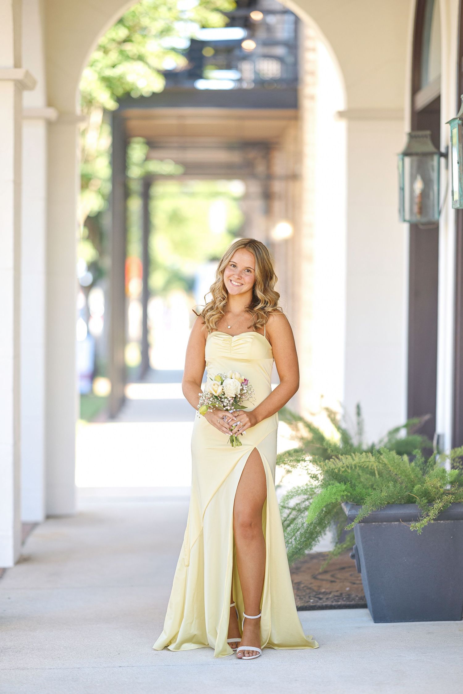 A figure in a pale yellow bridesmaid dress poses elegantly in a covered outdoor walkway with columns and planters.
