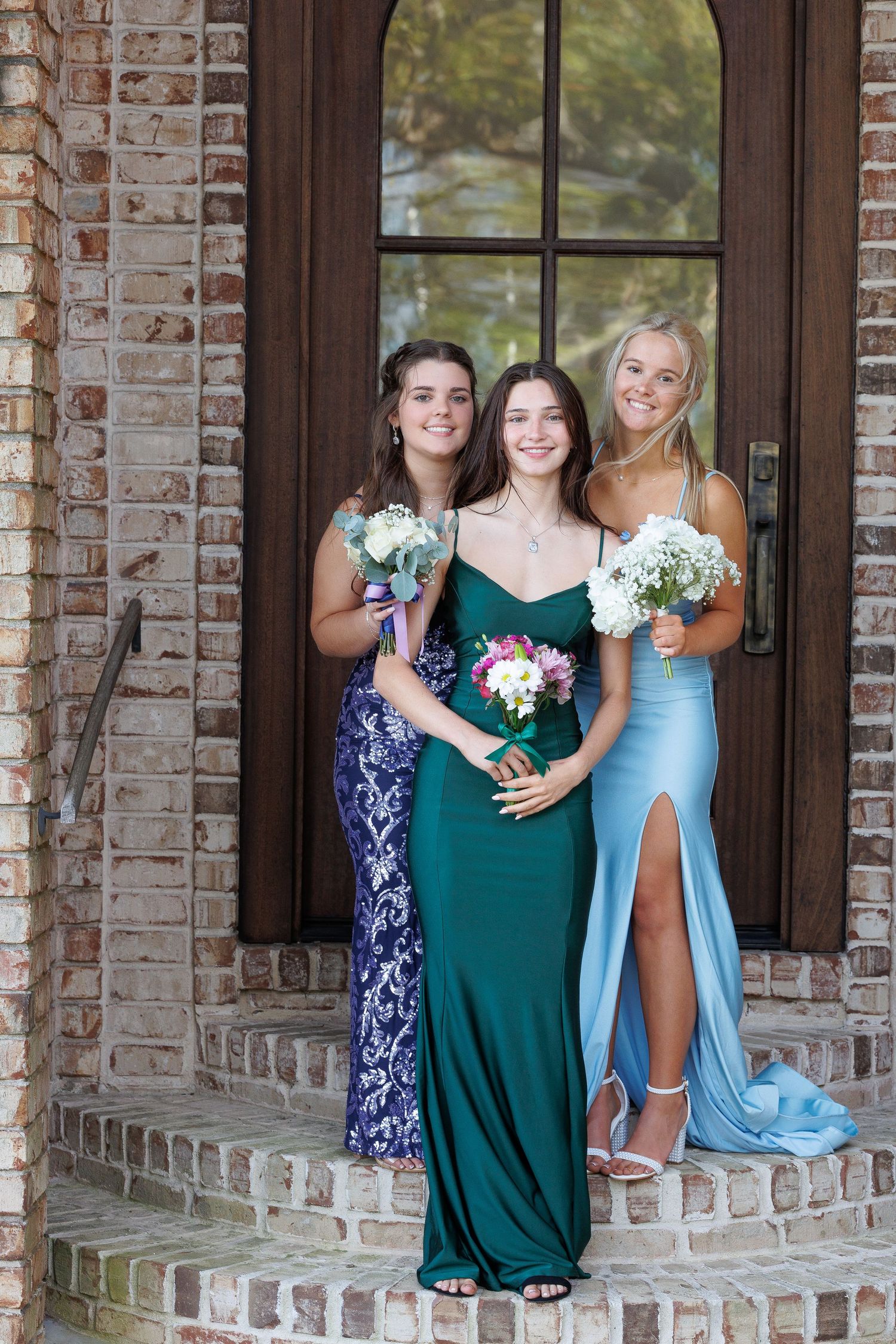 Three women in formal dresses stand together on brick steps holding flower bouquets in navy, emerald and light blue gowns.
