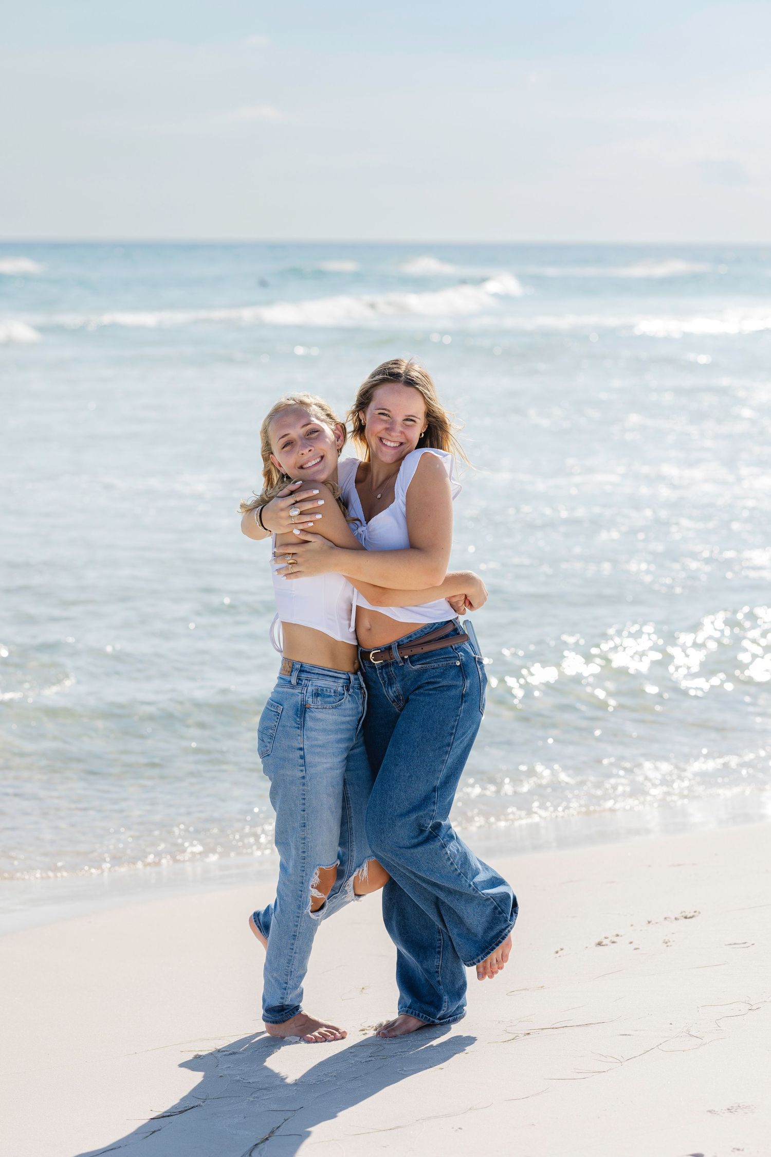 Two friends embracing and laughing together on a sunny beach with ocean waves in the background.