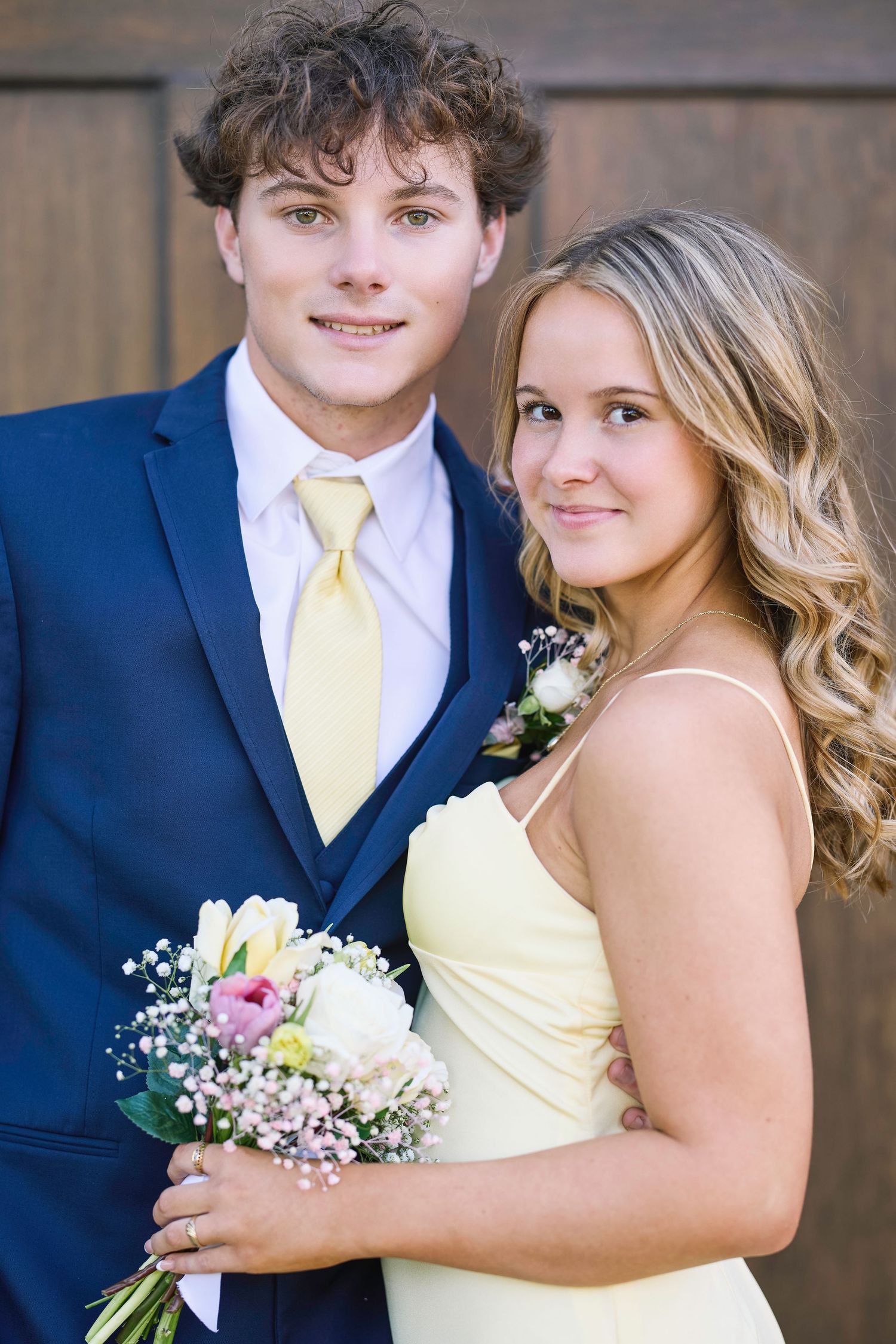 A couple in navy and pale yellow formal wear poses together with a small floral bouquet.
