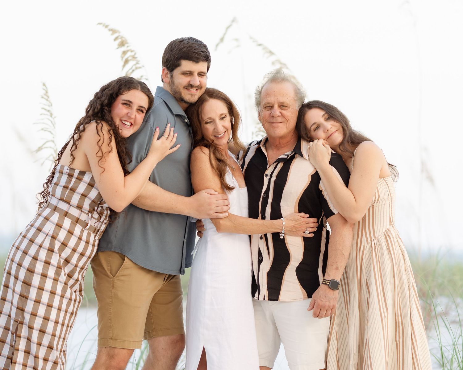 Happy family of five posing together outdoors at a beach, smiling and embracing near sea oats on a bright day.