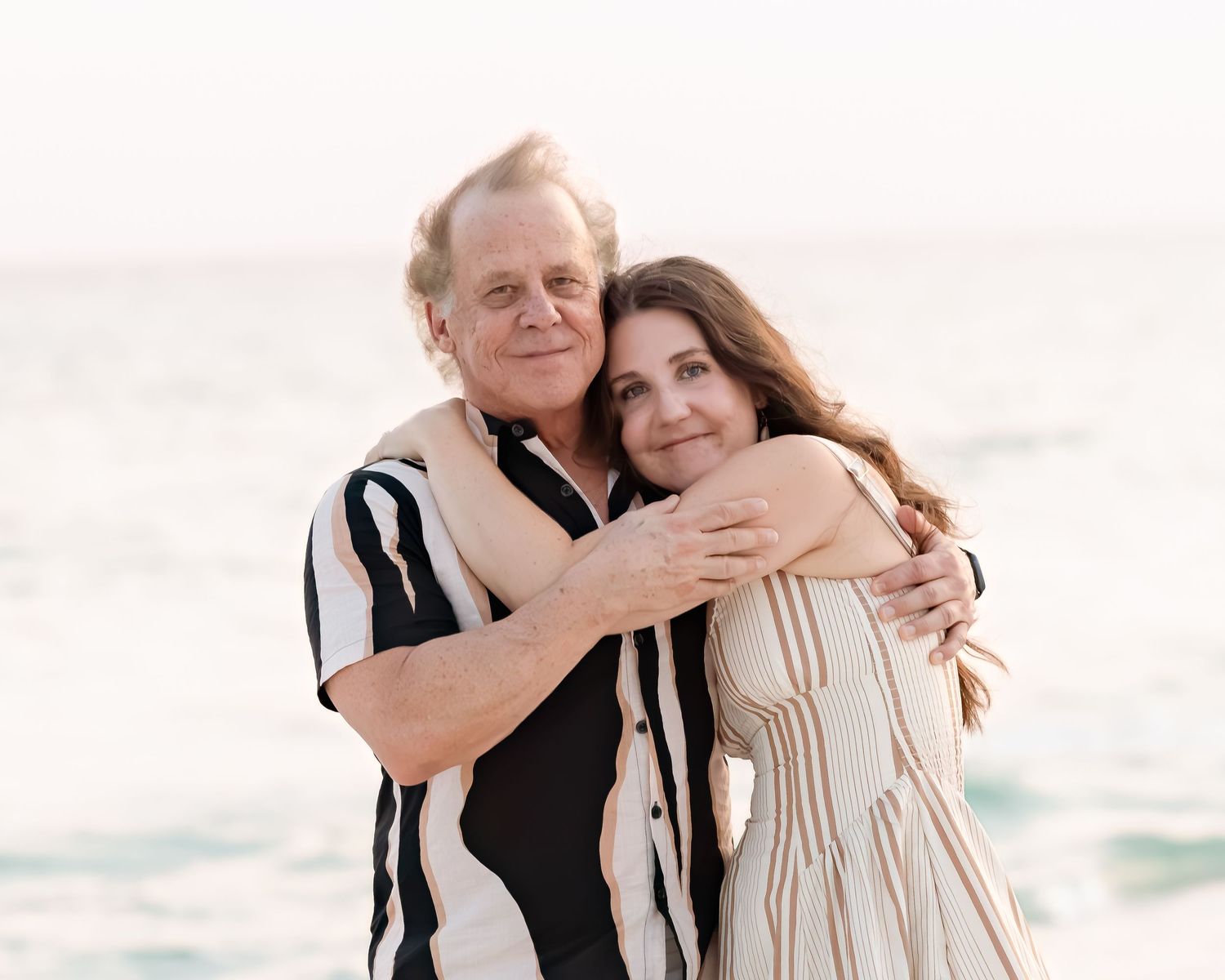 Older couple embracing on a beach, smiling warmly with ocean waves in the background.
