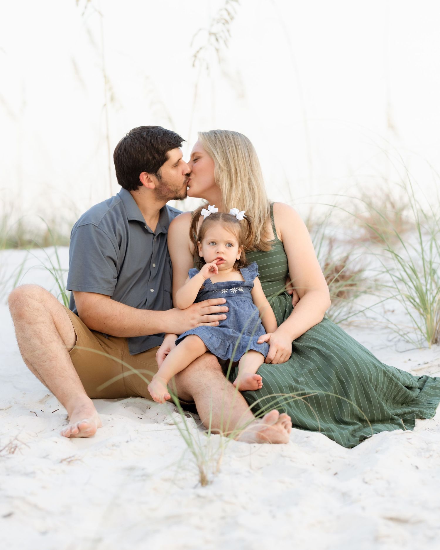A couple kisses on a white sand beach while their toddler daughter sits between them in a sweet family portrait.