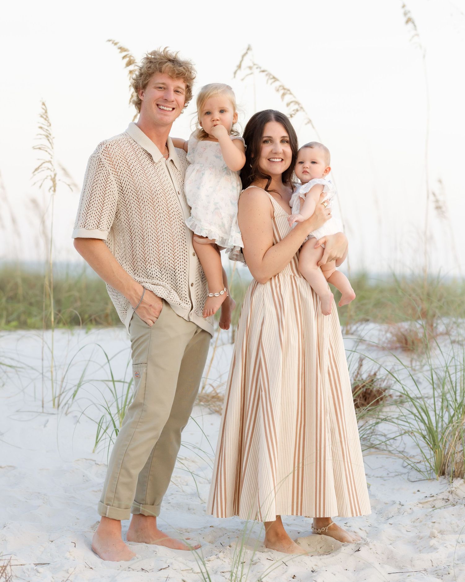 A young family of four poses on a sandy beach, parents holding two small children, dressed in neutral tones.