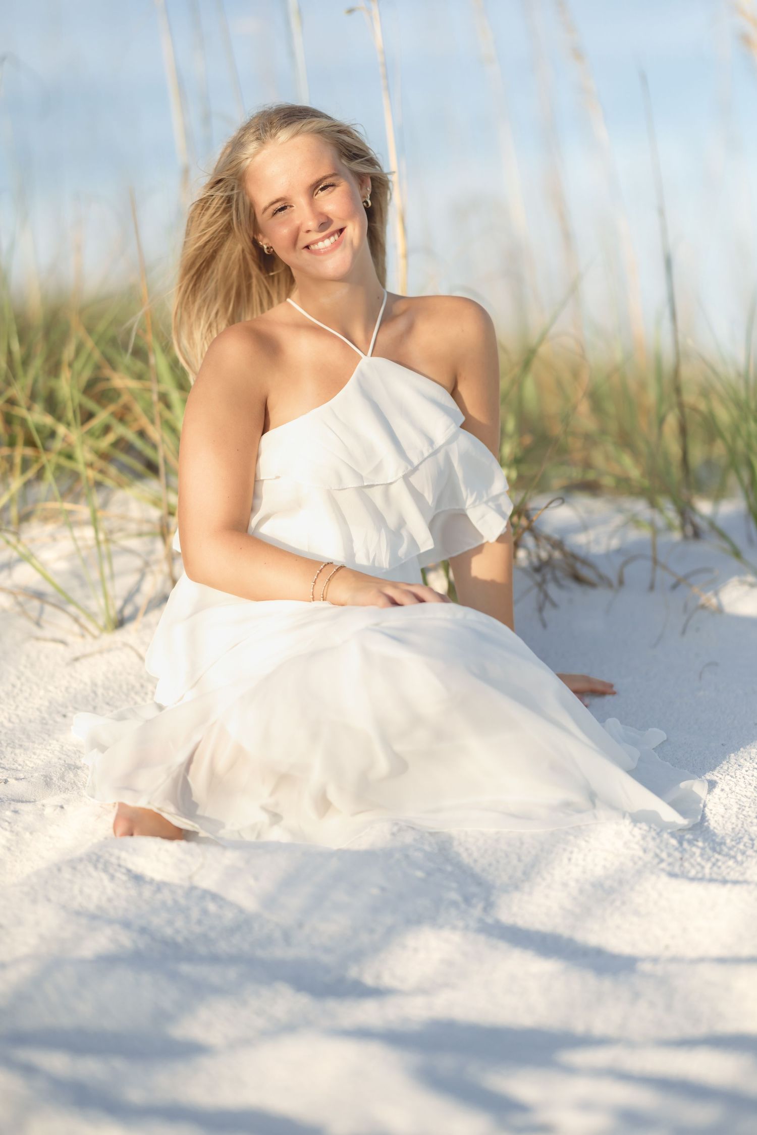 Young blonde woman in white dress sitting on white sand dunes with sea oats, smiling outdoors.