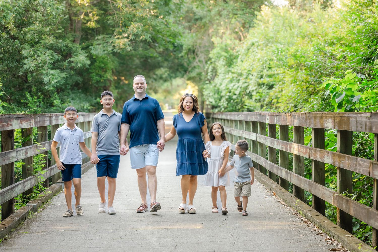 Family of six walking on a wooden bridge surrounded by lush green trees on a sunny day.