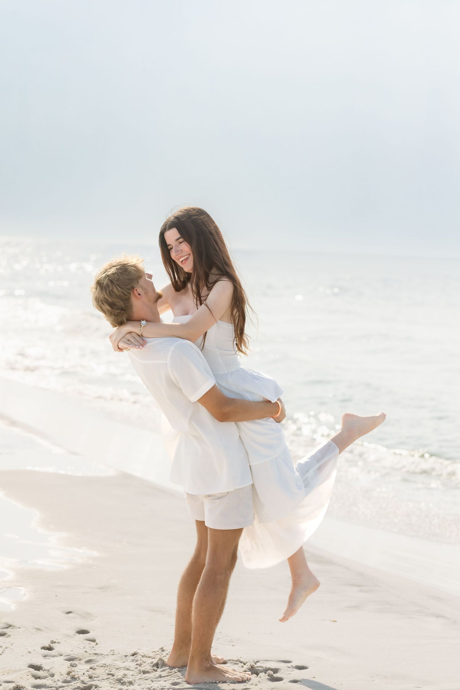 A couple in white outfits embracing on a sunny beach, man lifting woman as waves crash behind them.