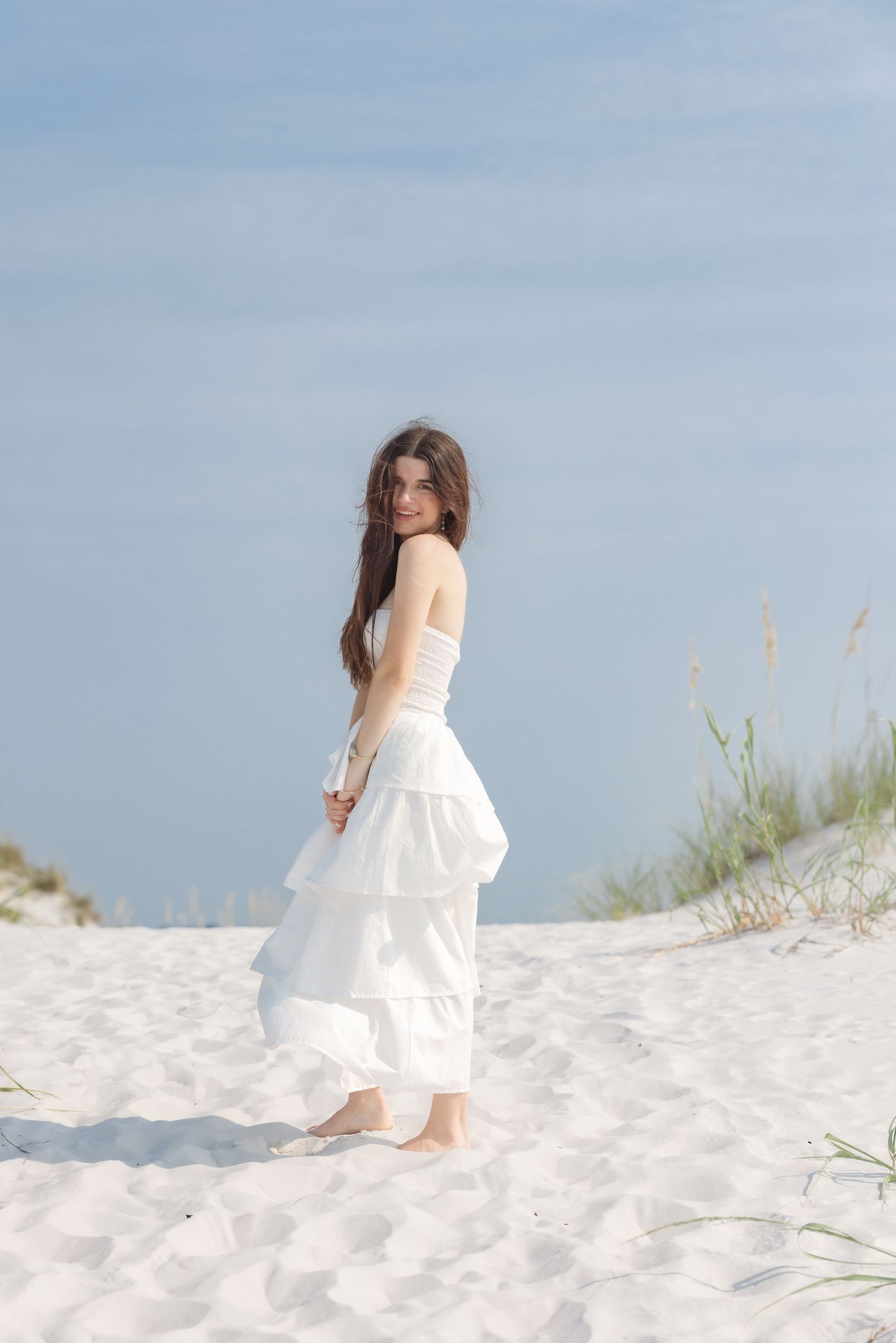Young woman in white tiered dress looking back over shoulder on sunny white sand dunes.