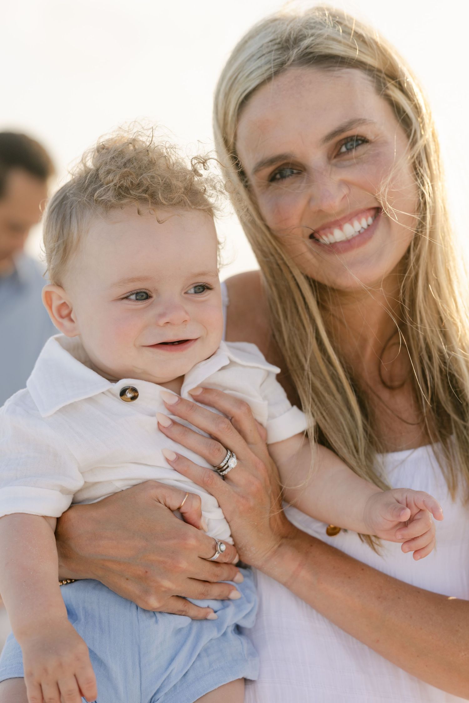 Smiling blonde woman holding a happy baby boy in white outfit at the beach on a sunny day.