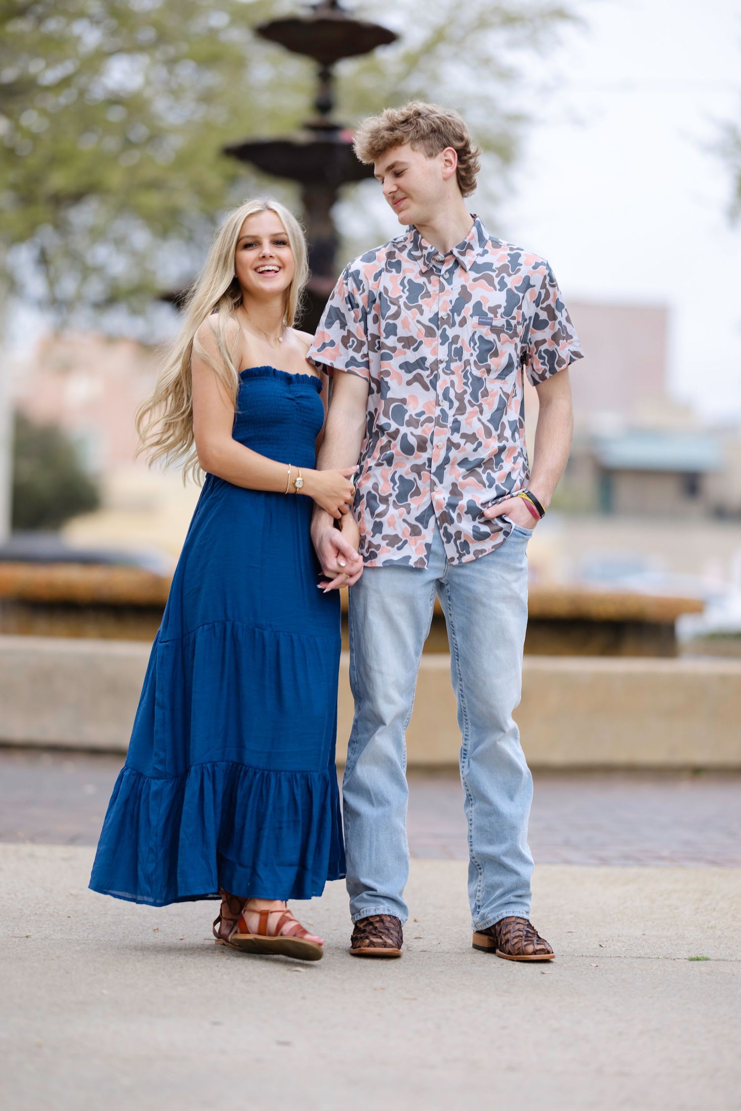 A couple poses together by a fountain, with her in a blue dress and him wearing a floral shirt and jeans.