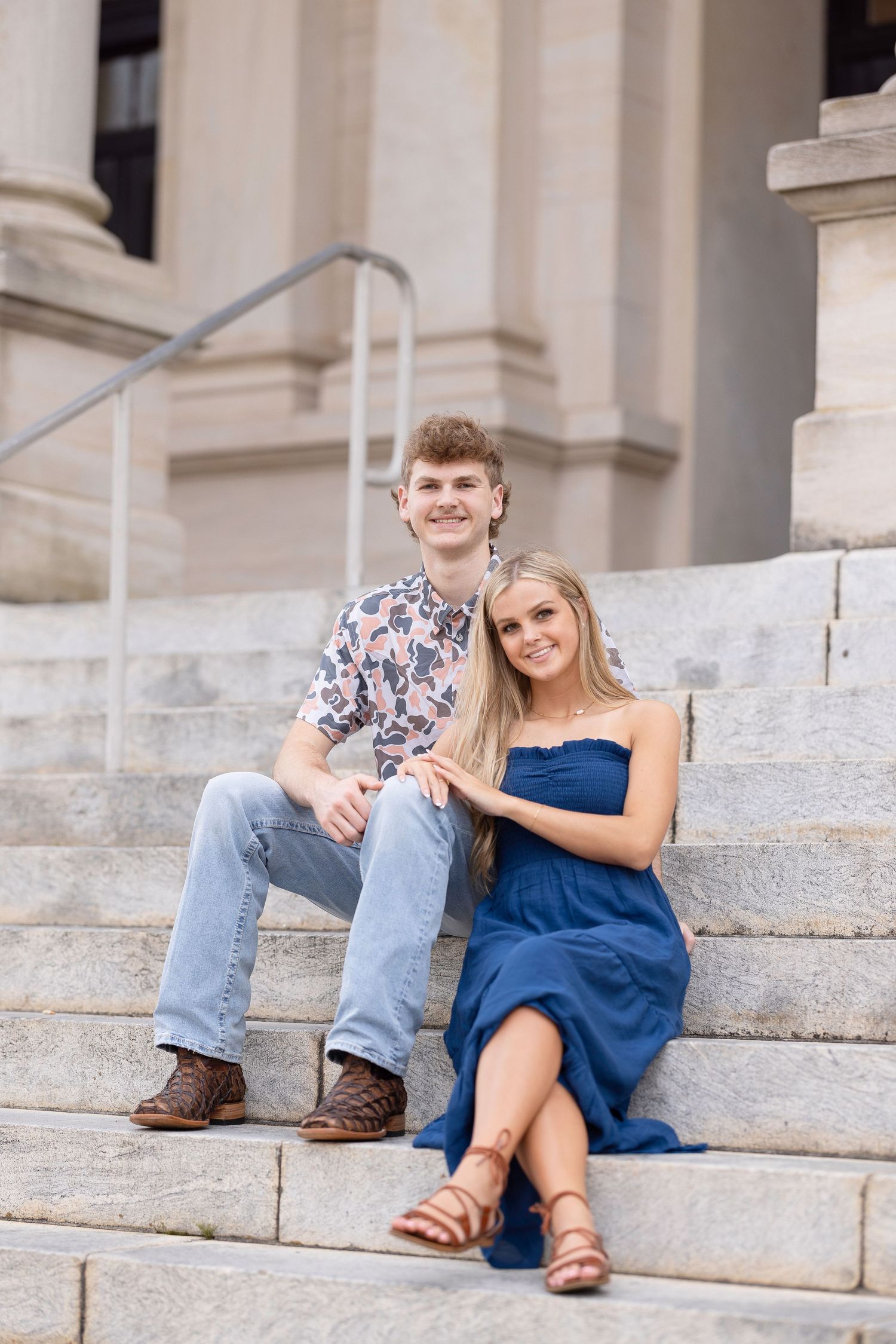 A couple sits together on white stone steps, with her wearing a blue dress and him in jeans and boots.
