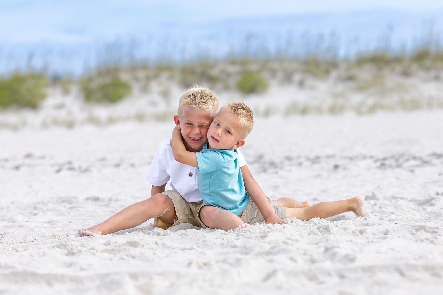 Two young boys hugging and playing together on a white sandy beach on a sunny day.