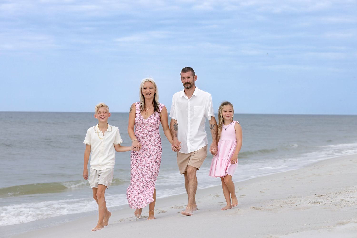 A happy family of four walks barefoot on a sunny beach, wearing coordinated pink and white outfits.