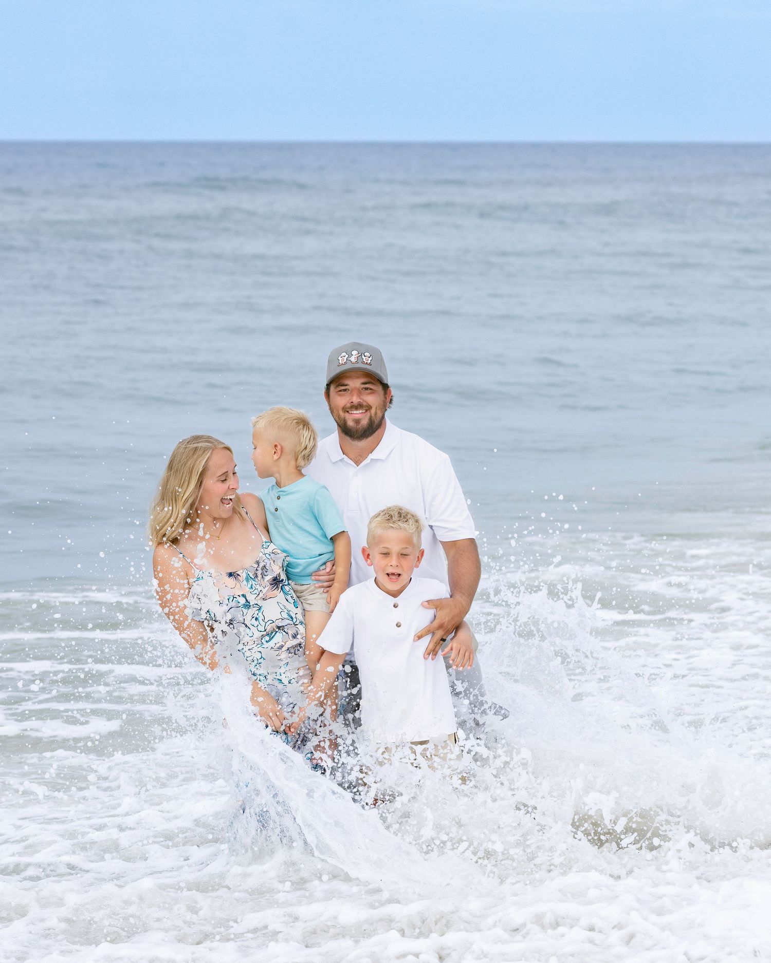 Family of four laughing and playing in ocean waves on a sunny beach day, parents holding young children.