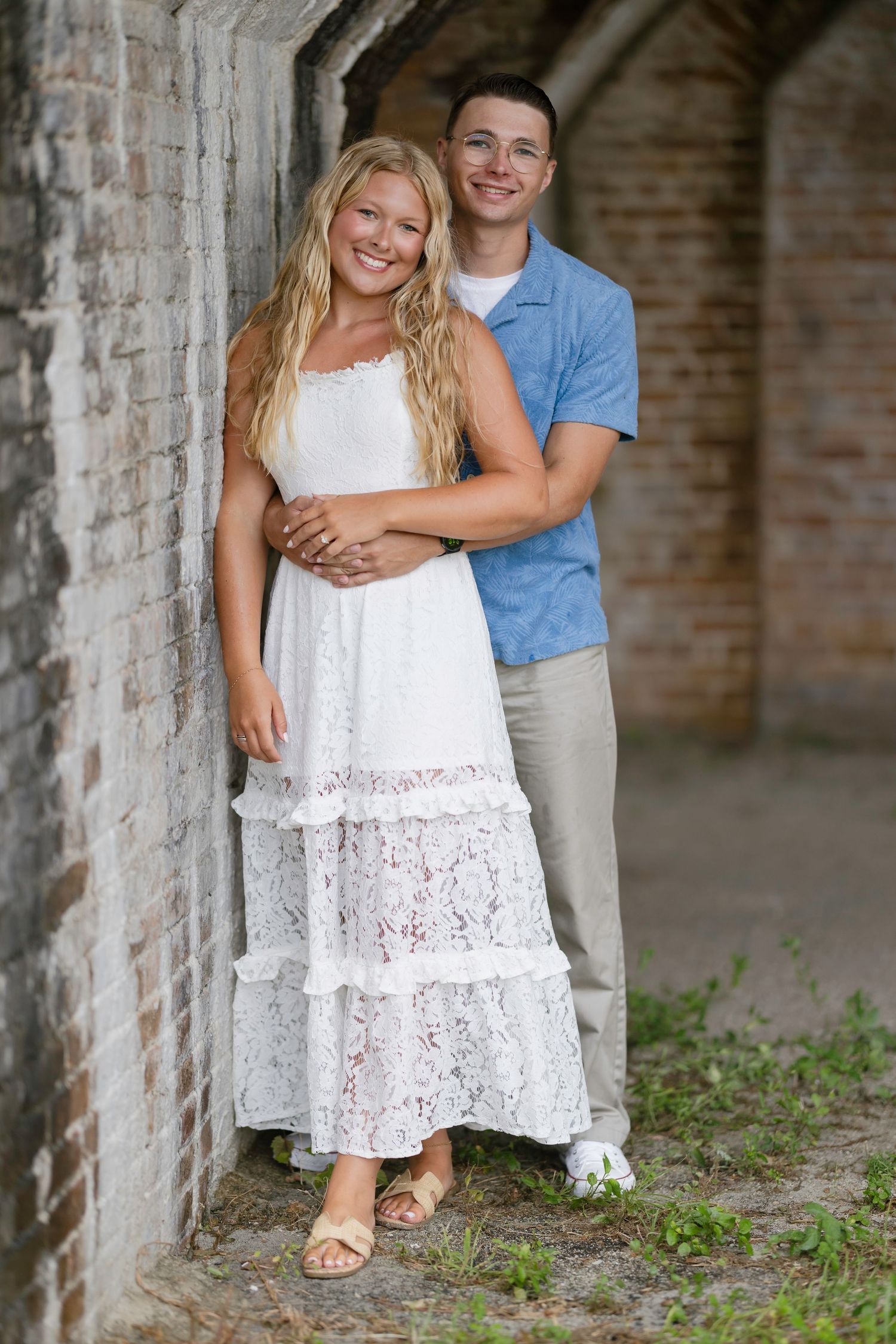 Young couple posing together, woman in white lace dress, man in blue shirt, leaning against brick wall outdoors.